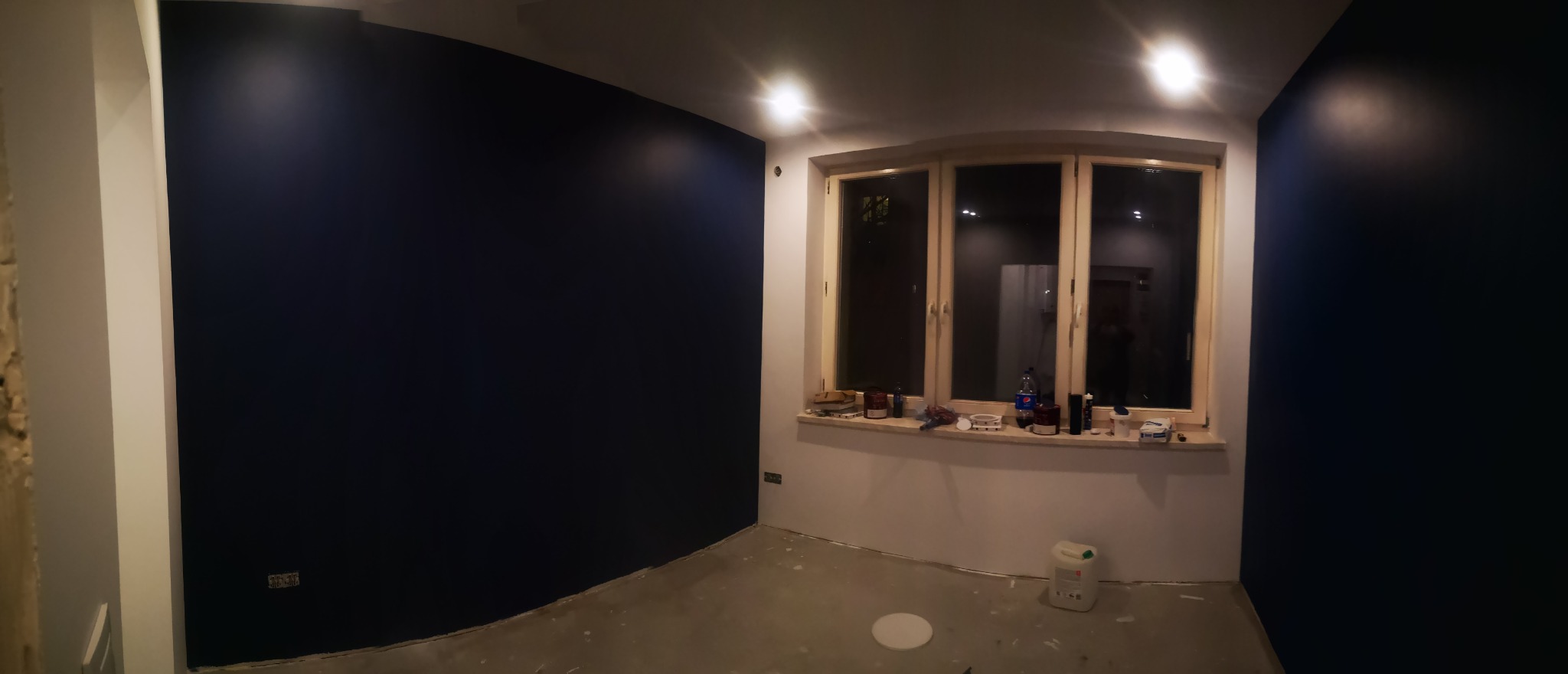 Pokój po malowaniu ścian na ciemny granat, widoczne niedoskonałości na podłodze i przy listwach, okno z akcesoriami malarskimi na parapecie.