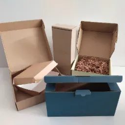 Różnorodne kartonowe pudełka otwarte i zamknięte, w tym jedno niebieskie z widocznym zamknięciem, wypełnione brązowym wypełniaczem, na białym tle.