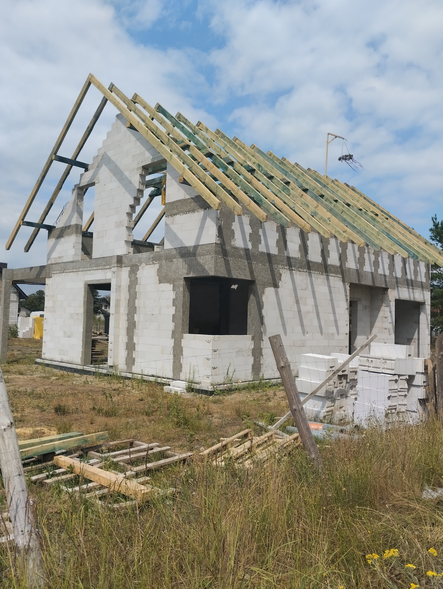 Surowy stan budynku z białych bloczków z zamontowaną drewnianą konstrukcją dachu, widok z poziomu gruntu, na pierwszym planie trawa i porozrzucane elementy drewniane.