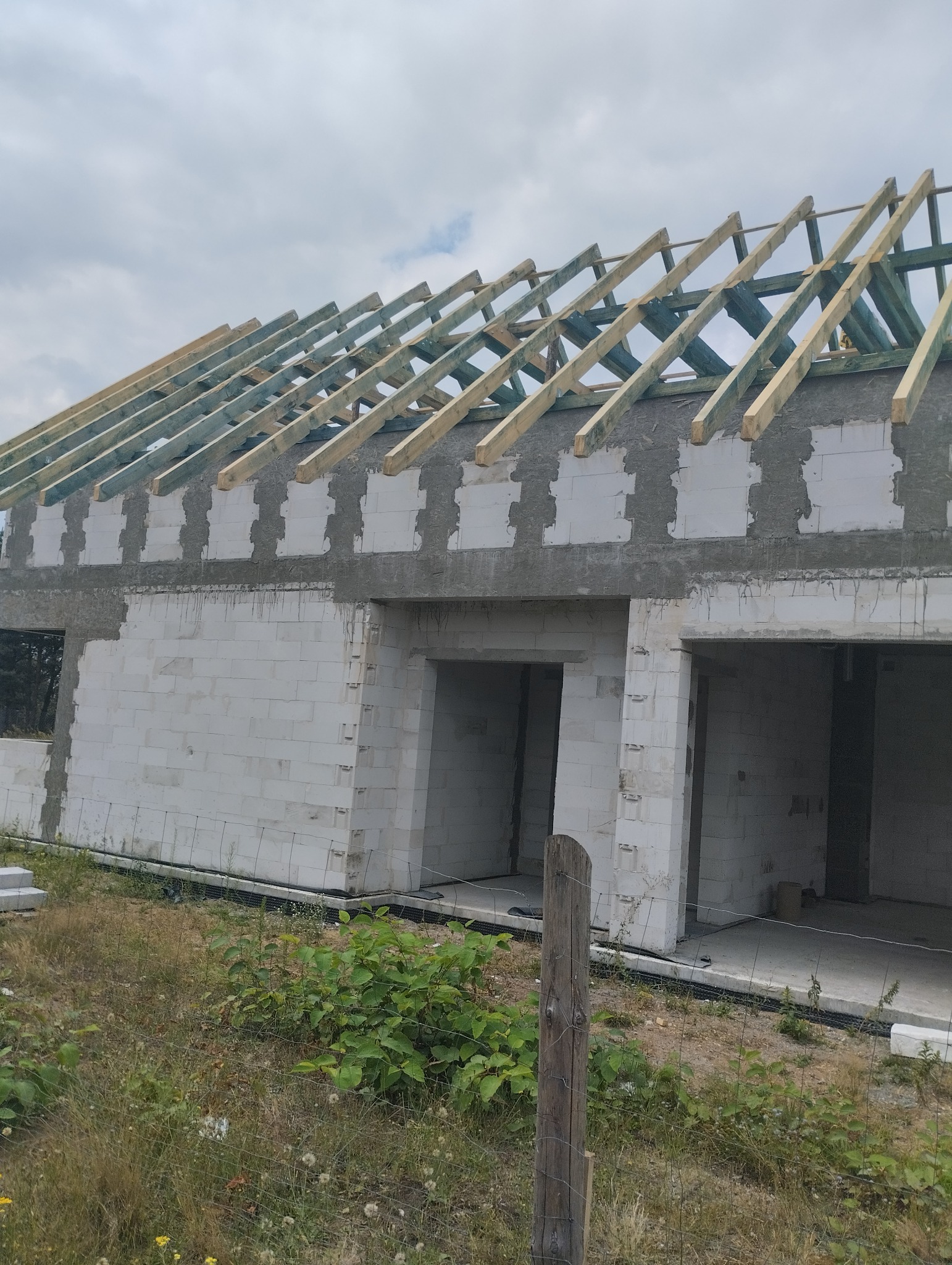 Surowy stan budowy domu z białego bloczka, widoczna drewniana konstrukcja dachu w trakcie montażu, zarośnięty teren wokół budynku.