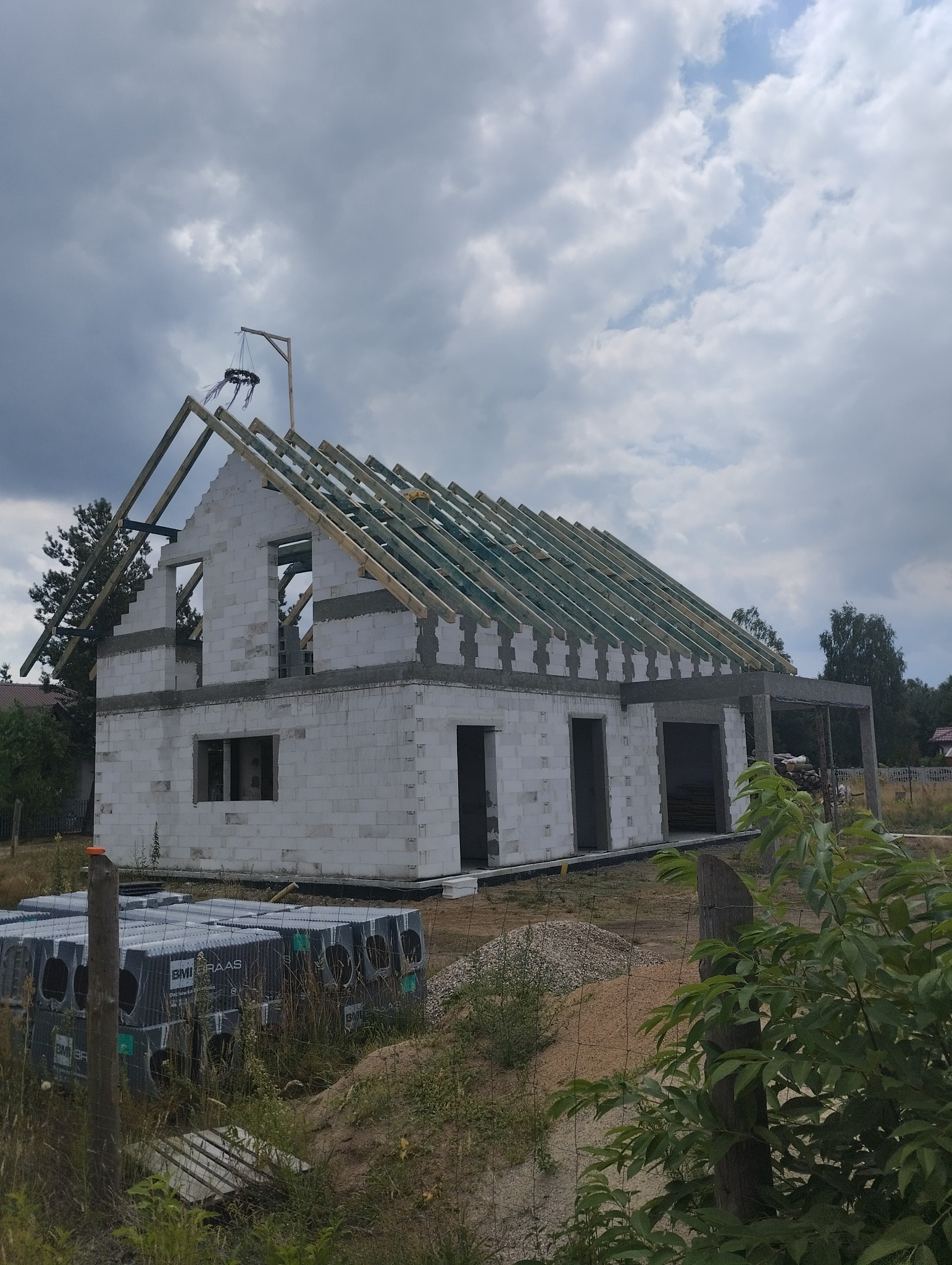 Budowa domu jednorodzinnego w stanie surowym otwartym: ściany z bloczków, drewniana konstrukcja dachu z widocznymi krokwiami i wieniec z betonu, na pierwszym planie palety z materiałami budowlanymi.