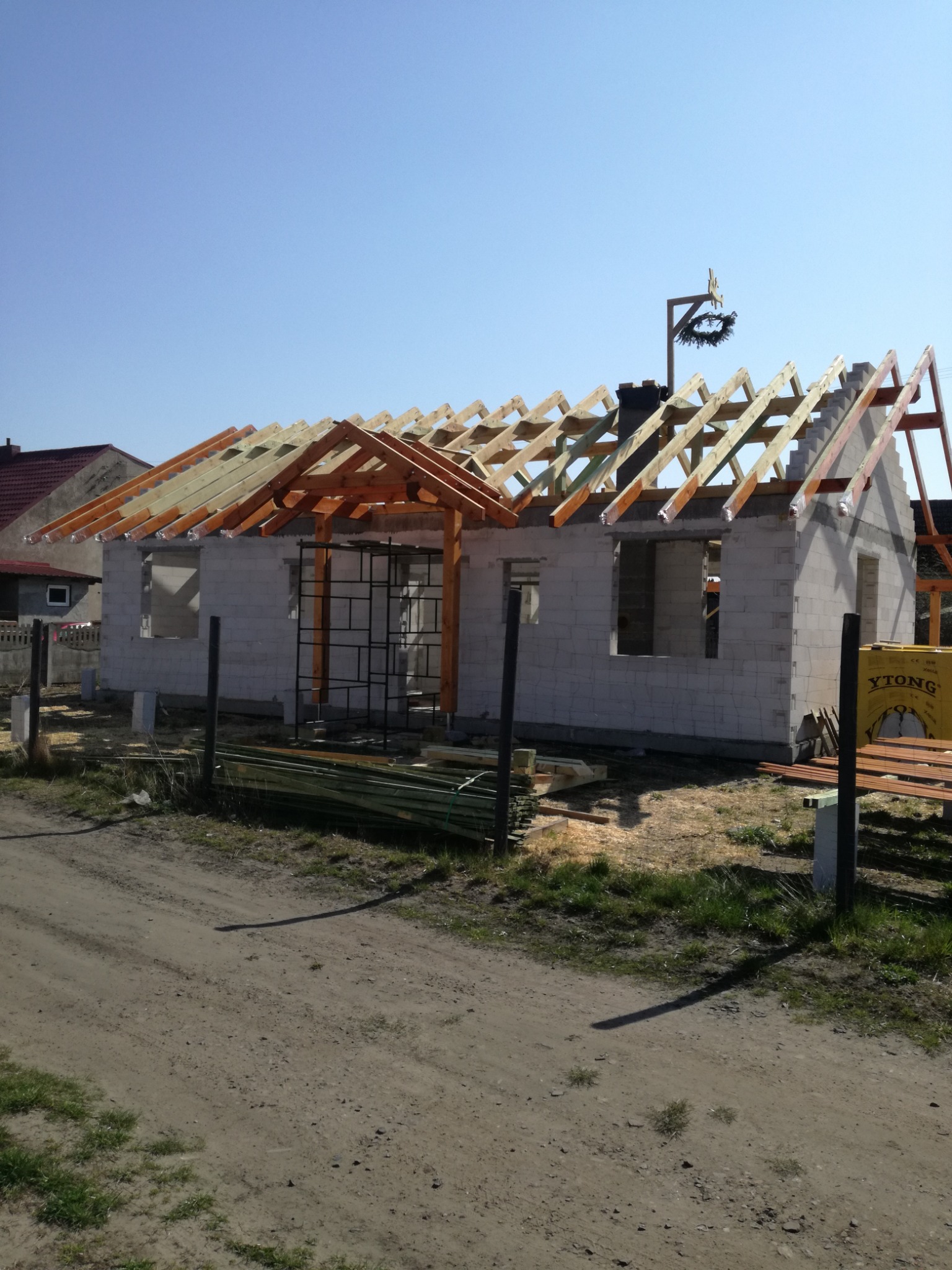 Surowy stan budowy domu z bloków Ytong, widoczna drewniana konstrukcja dachu z wieńcem na szczycie, rusztowanie metalowe przy ścianie, w tle zabudowa jednorodzinna.