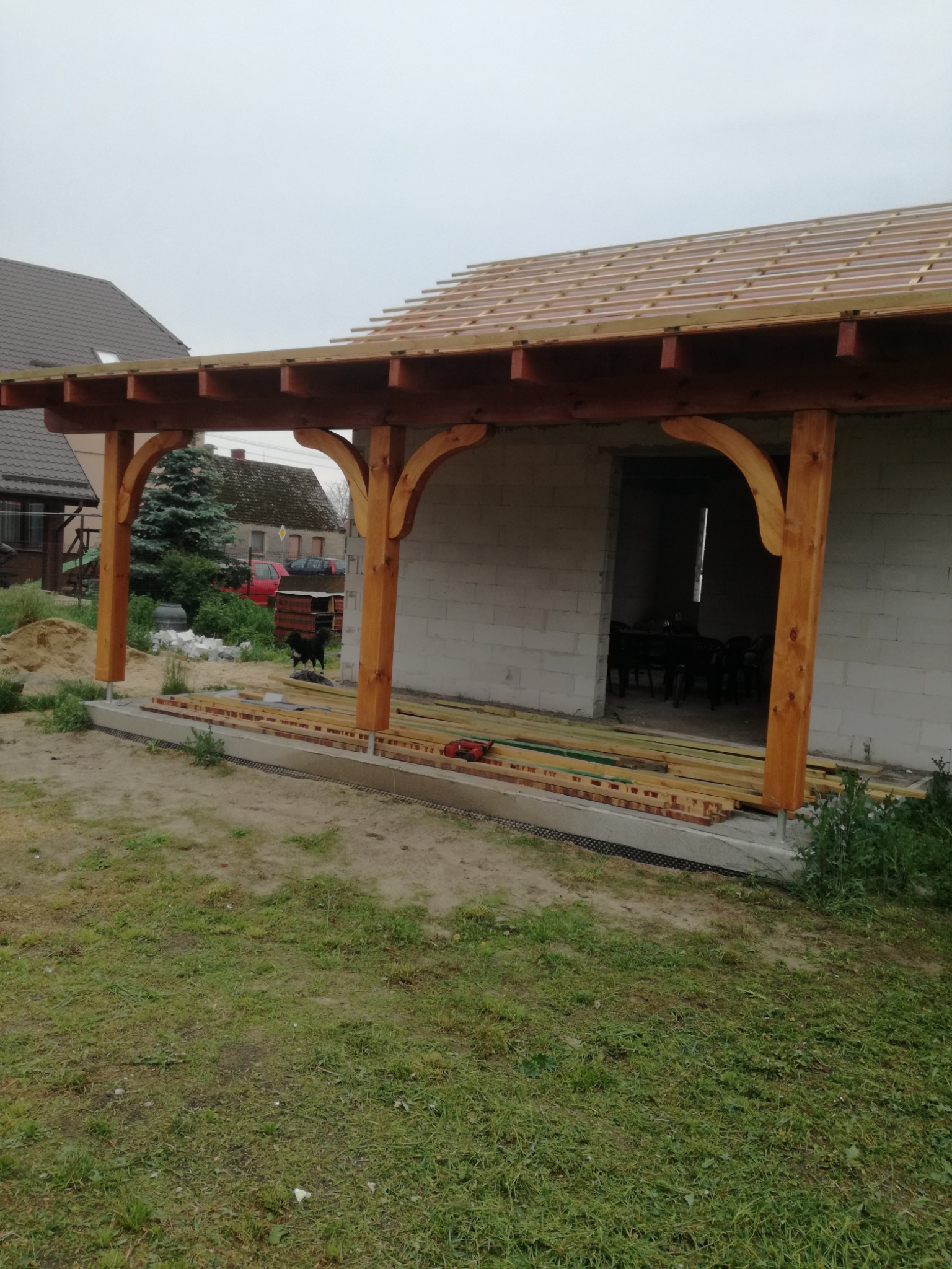 Drewniana konstrukcja z zaokrąglonymi podporami, prawdopodobnie w trakcie budowy, z widoczną więźbą dachową i elementami dachu w tle, na zewnątrz budynku z szarego bloczka.