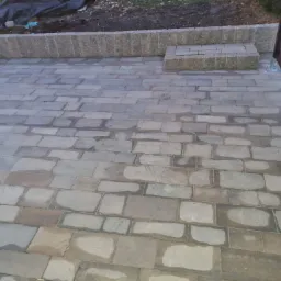 Ułożona szara kostka brukowa z widocznym fragmentem ogrodzenia z drewnianych desek i betonowym obrzeżem. Część kostki jest mokra.