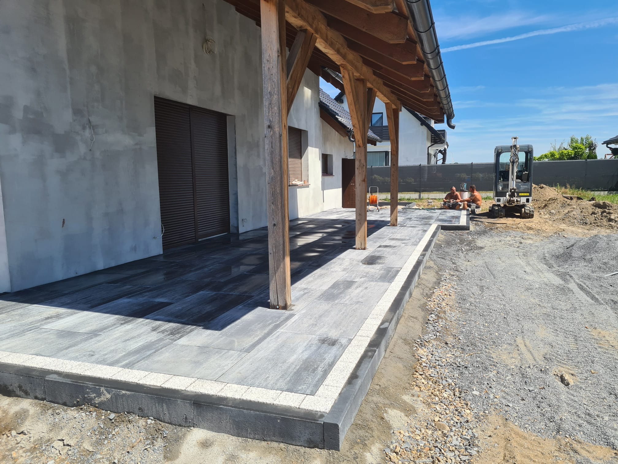 Nowoczesny taras z szarych płyt kamiennych, wykończony jasną kostką, z drewnianą konstrukcją zadaszenia i mini koparką w tle, dwóch robotników w trakcie przerwy.