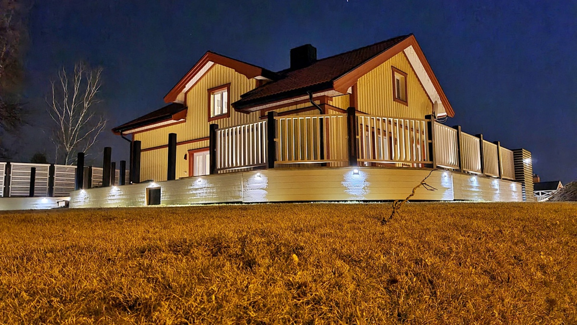 Oświetlony, dwukondygnacyjny dom z drewnianą elewacją w kolorze kremowym i czerwonym dachem, widoczny nocą z poziomu trawnika.
