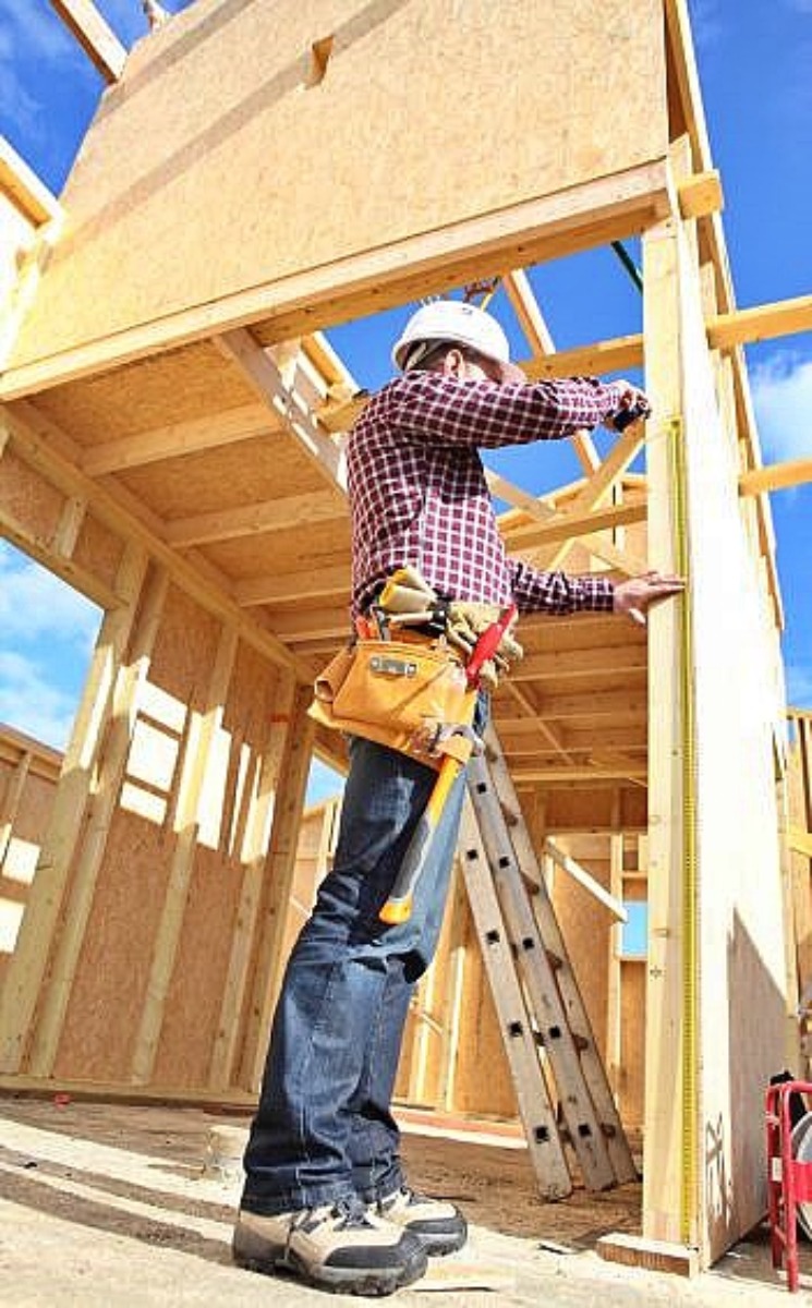 Pracownik w kasku mierzy ścianę w trakcie budowy drewnianej konstrukcji, widoczna drabina i pas z narzędziami.