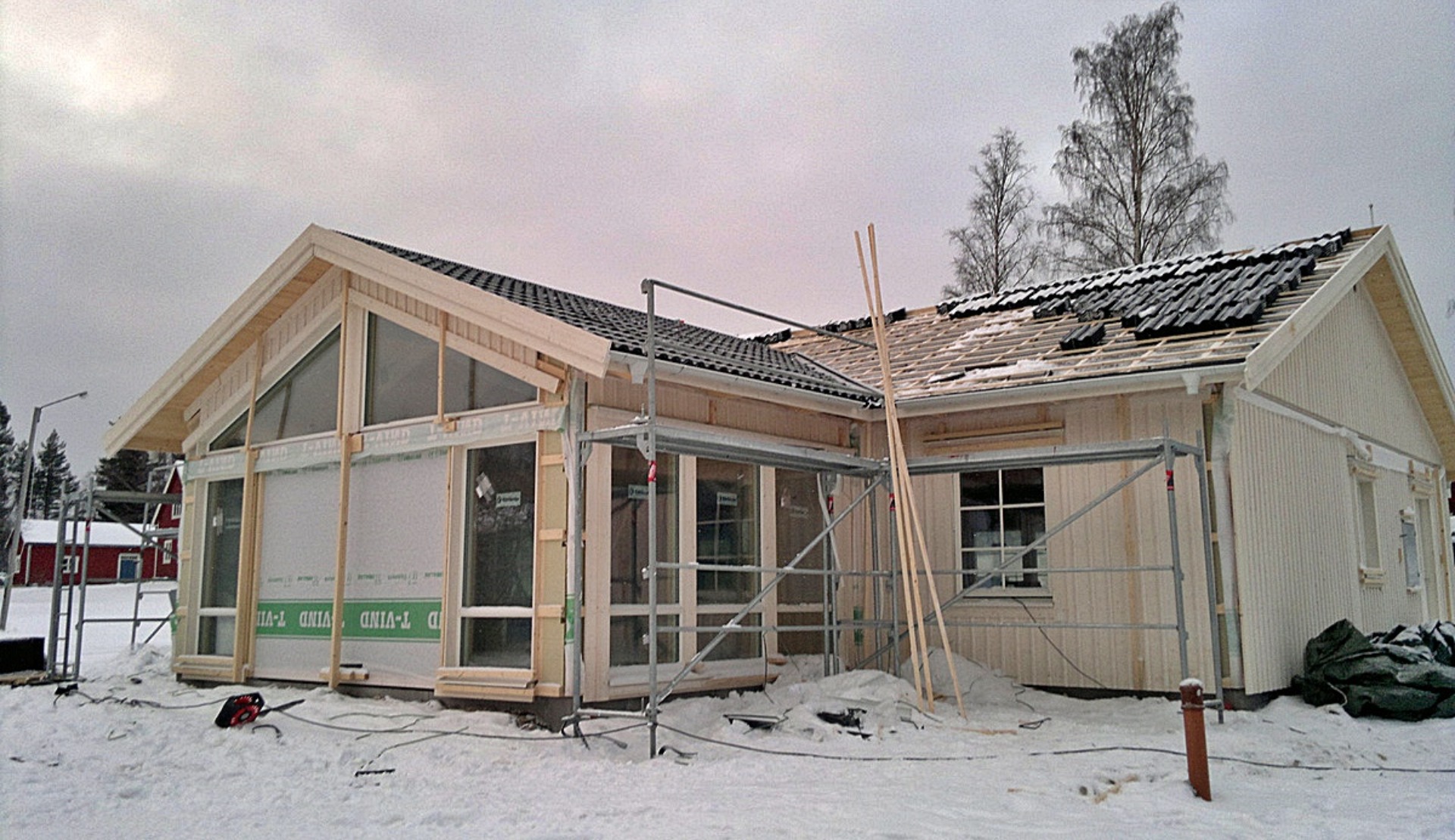 Surowy, drewniany dom w trakcie budowy zimą, z widocznymi rusztowaniami, częściowo ułożonym dachem z czarnej dachówki i zabezpieczonymi oknami.
