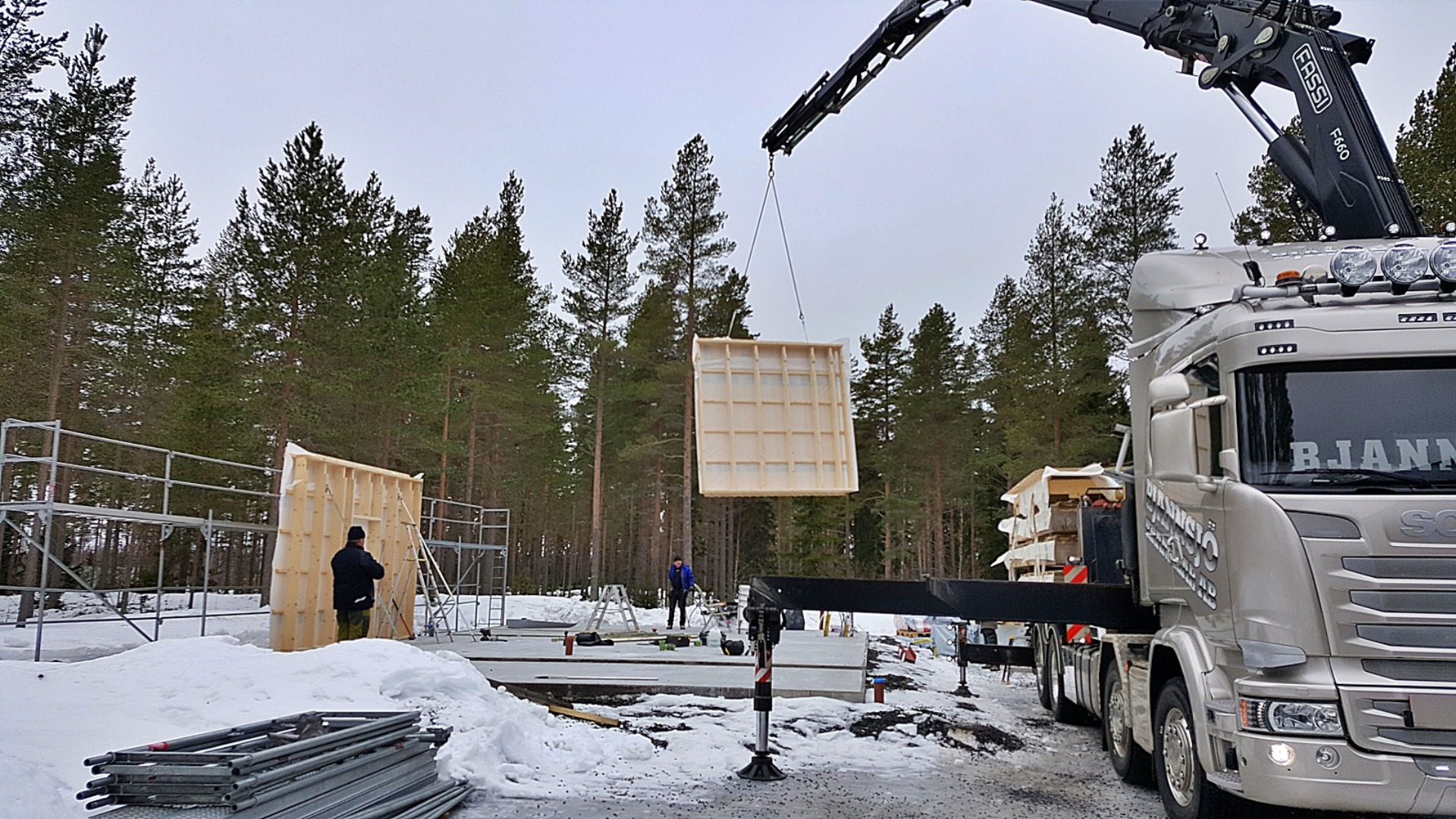Montaż ściany z drewnianych paneli prefabrykowanych za pomocą dźwigu w Szczecinie, widoczny fragment konstrukcji budynku na fundamencie, w tle las i śnieg.