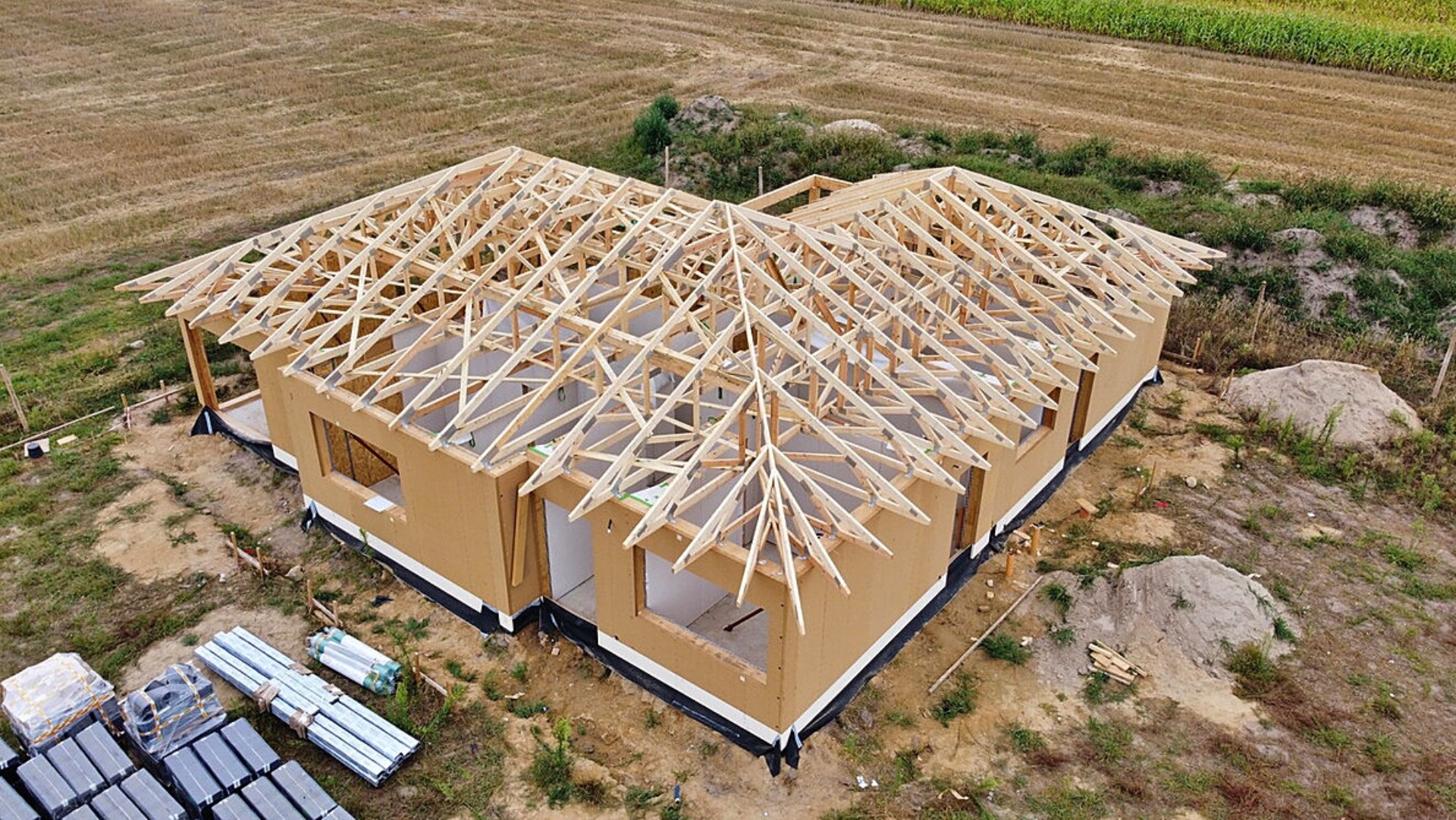 Wznoszenie szkieletu dachu na parterowym domu szkieletowym z widocznymi elementami konstrukcyjnymi, materiałami budowlanymi i przygotowanym terenem pod budowę.