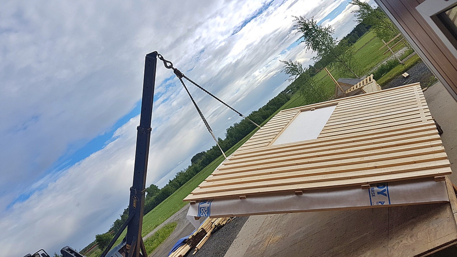 Panel ściany z oknem podnoszony żurawiem na placu budowy, widok z dołu na tle zieleni i pochmurnego nieba.