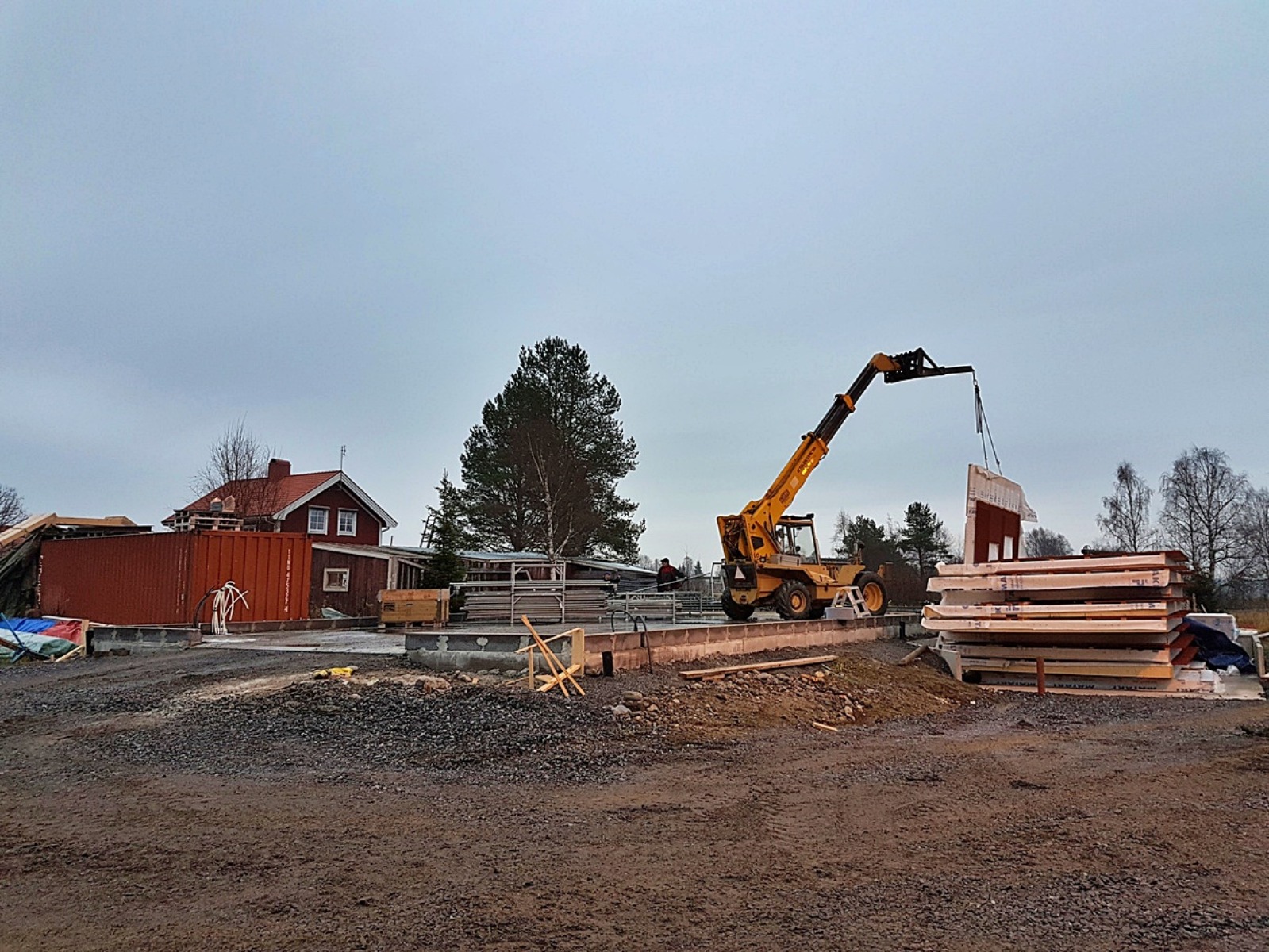 Żółta ładowarka teleskopowa podnosi gotowy panel ścienny na placu budowy fundamentów pod budynek, w tle czerwony budynek gospodarczy.