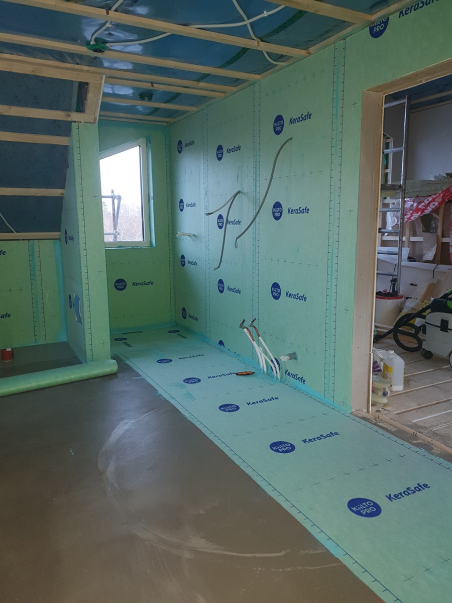 Wnętrze w trakcie budowy z zielonymi płytami KeraSafe na ścianach, widoczne przewody instalacji elektrycznej i wodnej, surowy beton na podłodze i drewniana konstrukcja stropu.