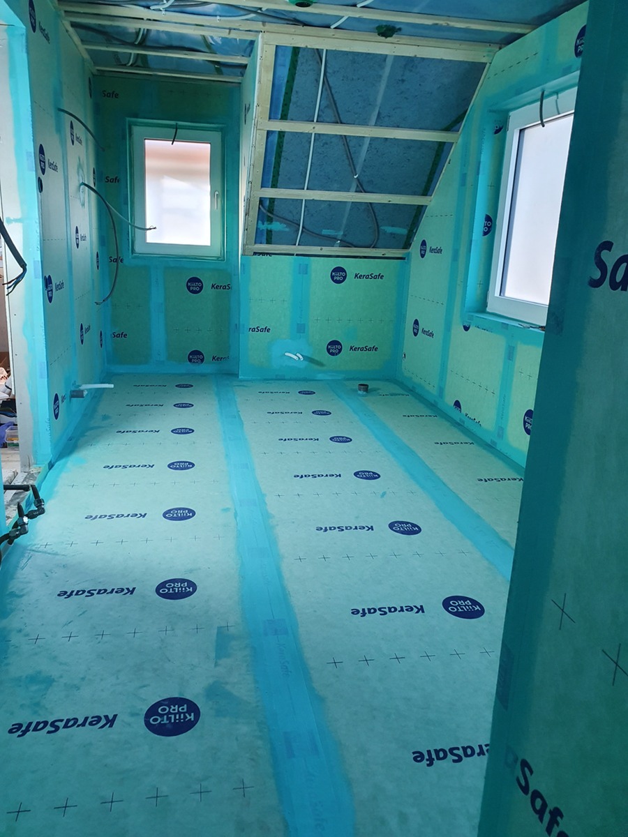 Wnętrze domu w trakcie budowy, widoczne ściany i podłoga zabezpieczone folią paroizolacyjną z nadrukiem KeraSafe, drewniana konstrukcja dachu i okna w ścianach szczytowych.