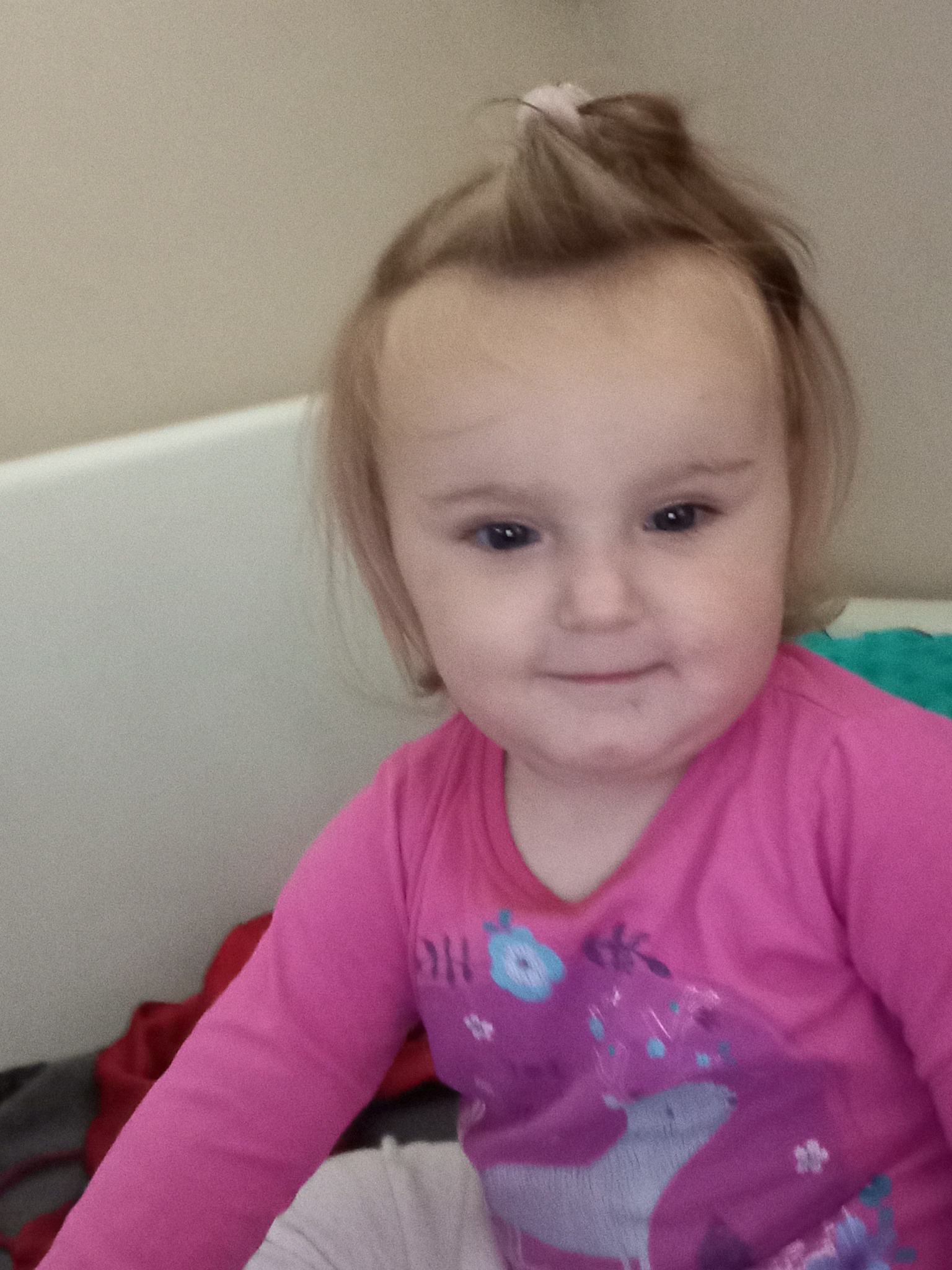 Portret uśmiechniętej dziewczynki w różowej bluzce z nadrukiem jednorożca, z włosami spiętymi w mały kucyk na czubku głowy, na tle jasnej ściany i pościeli.