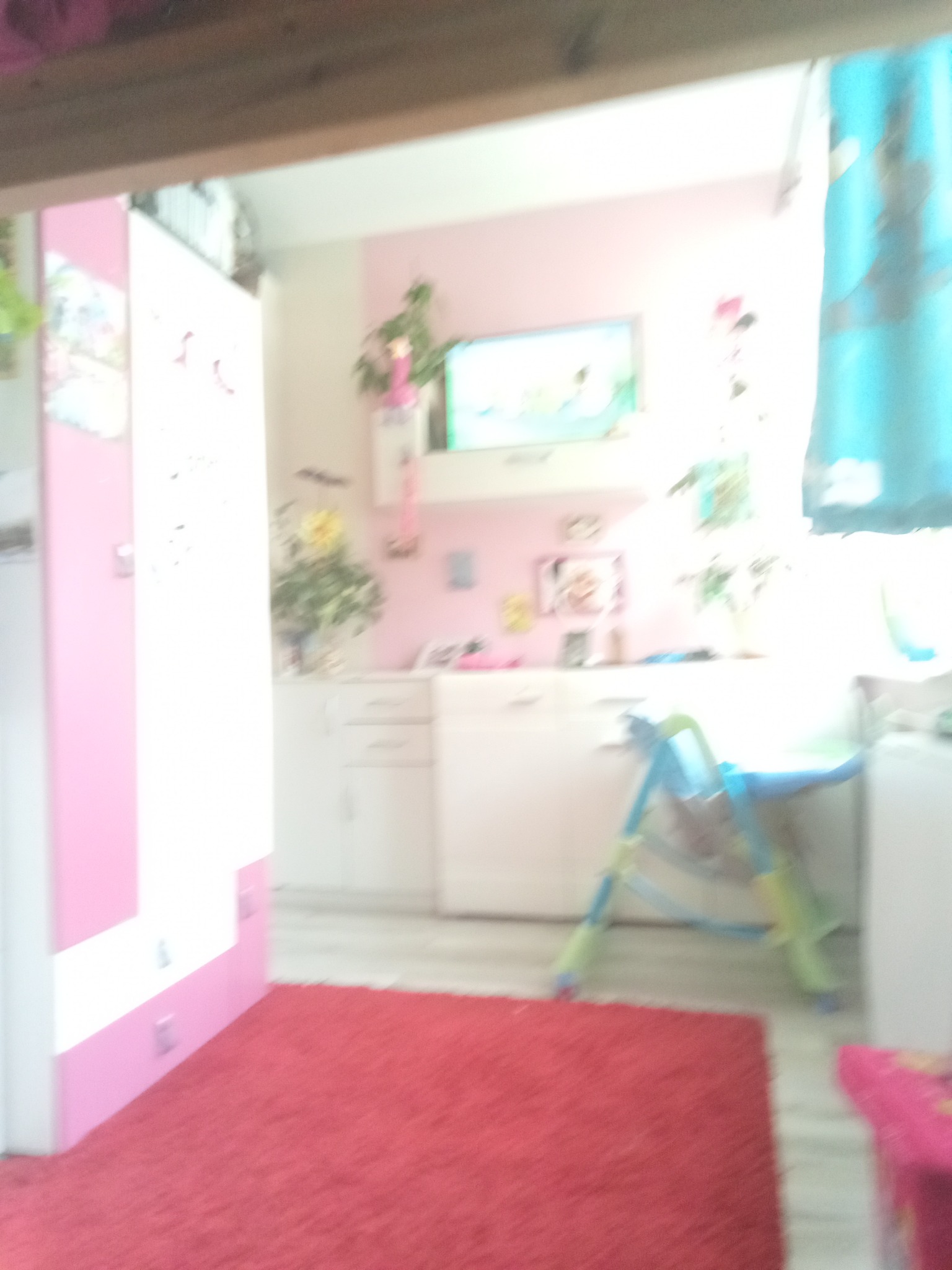 Pokój dziecięcy po odświeżeniu, z różowymi akcentami na ścianach i meblach, widoczny czerwony dywan i dziecięce krzesełko do karmienia w tle, całość w pastelowych barwach.