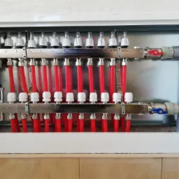 Rozdzielacz ogrzewania podłogowego z czerwonymi rurami w metalowej szafce, widoczne białe regulatory przepływu i napisy odręczne na belce rozdzielacza.