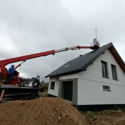 Montaż instalacji odgromowej na dachu domu jednorodzinnego z użyciem podnośnika koszowego na samochodzie ciężarowym, widoczny stos ziemi.