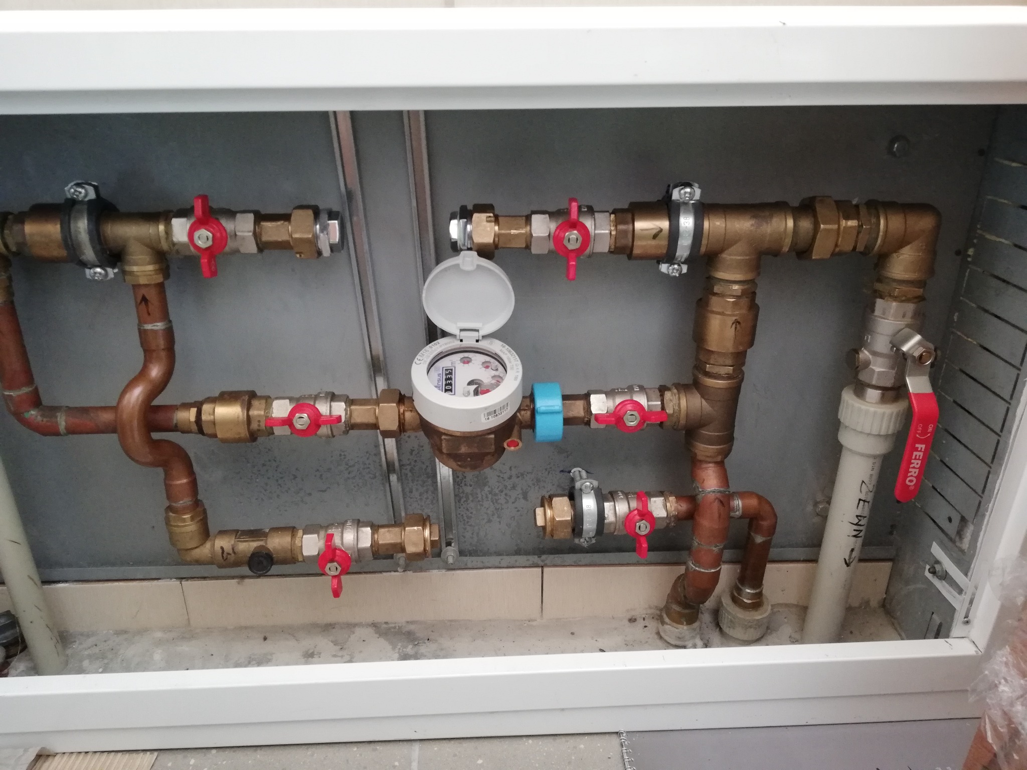 Zabudowany licznik wody z otwartą klapką, widoczne miedziane i mosiężne rury, zawory z czerwonymi pokrętłami, instalacja wodna wewnątrz szafki, szare tło.
