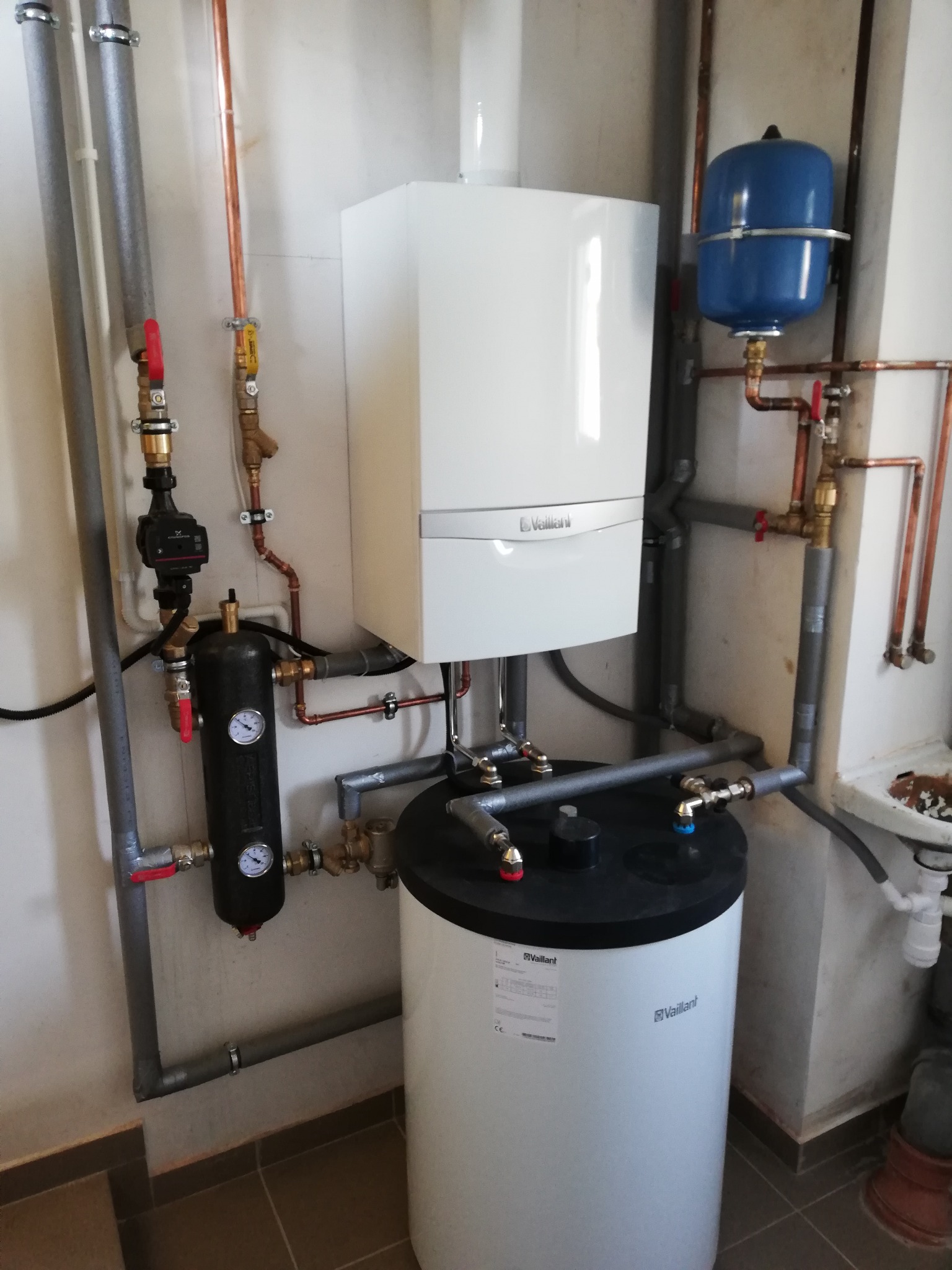 Biały piec gazowy Vaillant z podłączonym zasobnikiem ciepłej wody, instalacją hydrauliczną z rur miedzianych i szarych, manometry na czarnym zbiorniku, niebieski zbiornik wyrównawczy, widoczne...