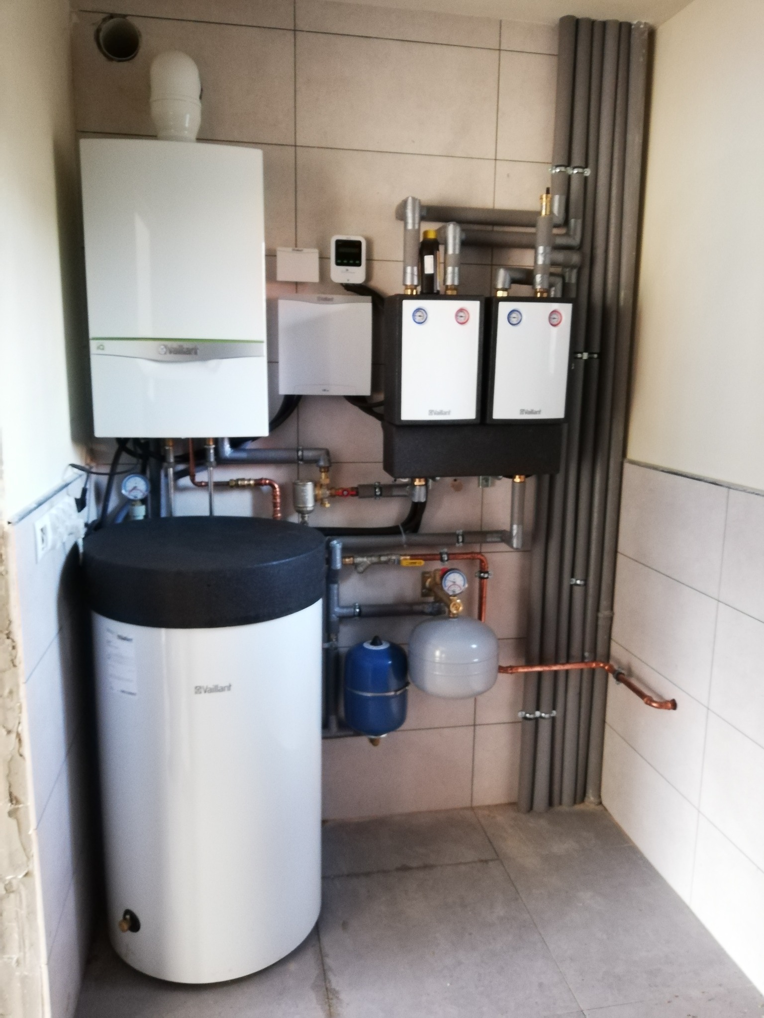 Kompletna instalacja grzewcza z kotłem gazowym, zasobnikiem ciepłej wody, dwoma obiegami grzewczymi i osprzętem marki Vaillant, zamontowana na kafelkowanej ścianie.