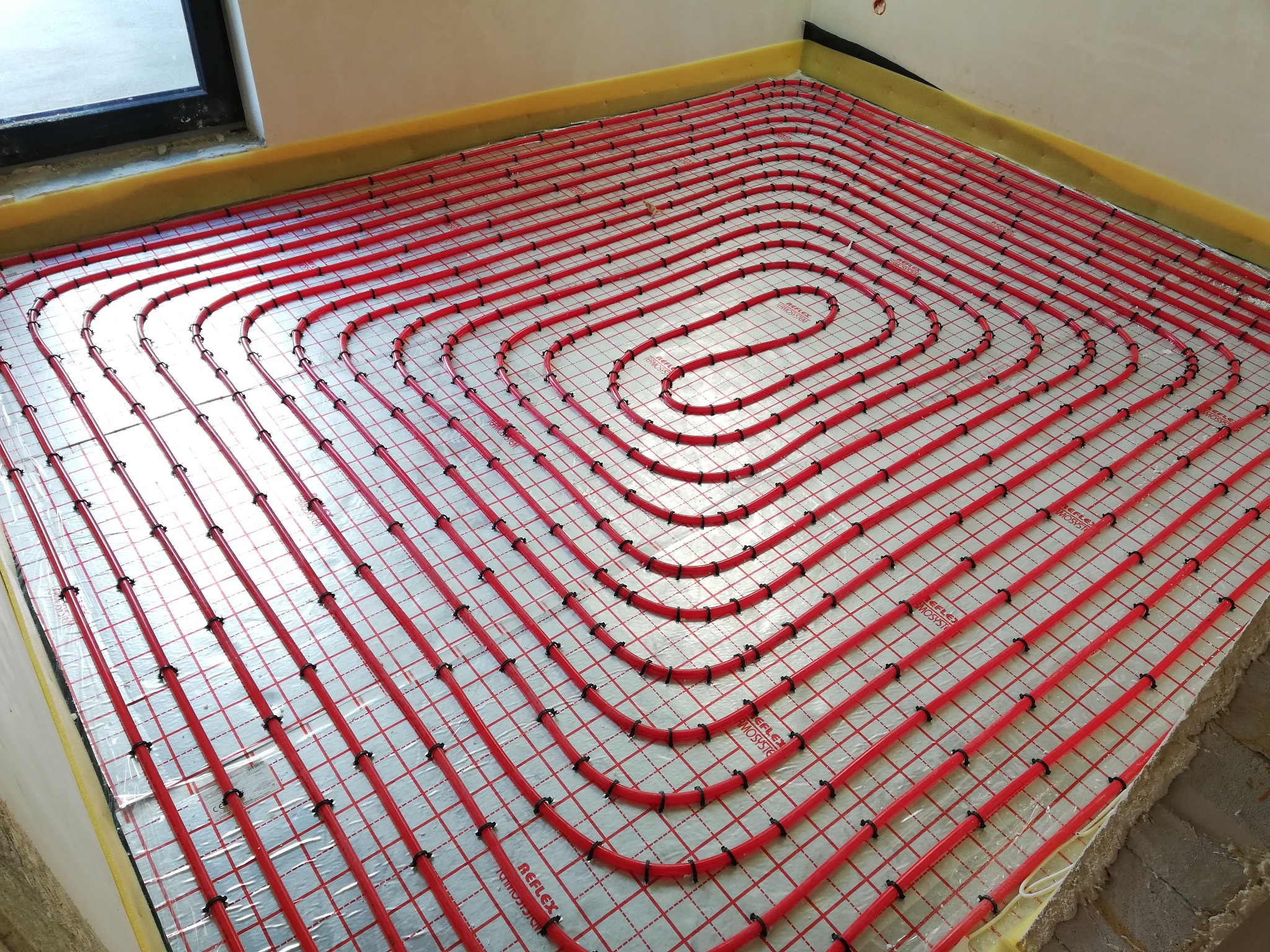 Czerwone rury ogrzewania podłogowego ułożone spiralnie na srebrnej folii z czerwoną siatką, przymocowane czarnymi klipsami, widoczne żółte paski izolacyjne przy ścianach i fragment okna w tle.