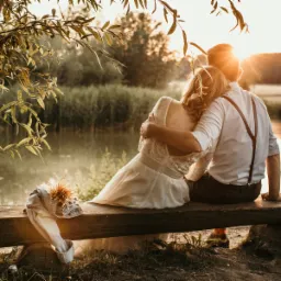 Para narzeczonych siedzi na drewnianej ławce nad jeziorem o zachodzie słońca, buty i bukiet leżą obok nich.