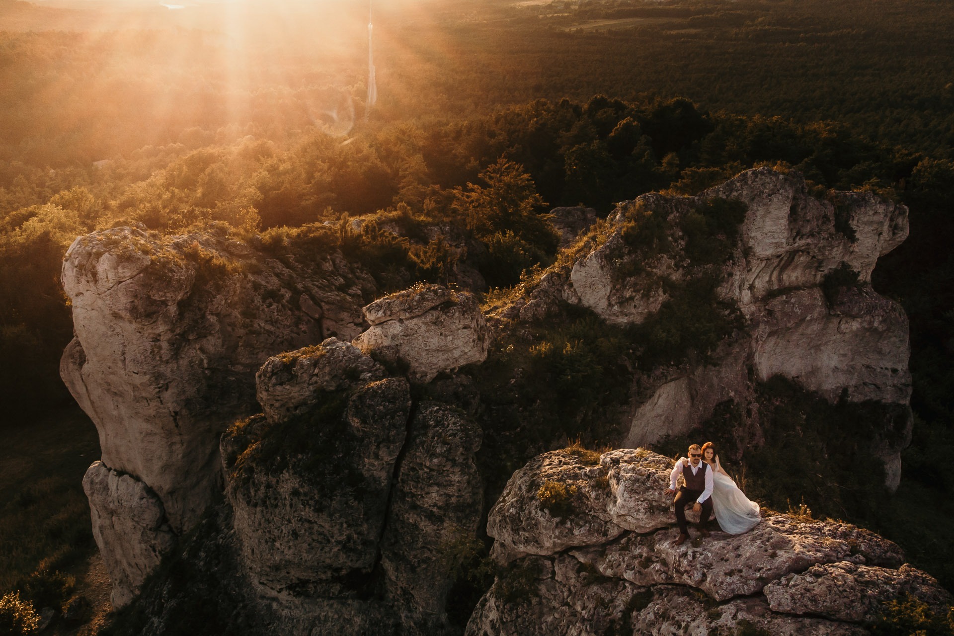 Para w strojach ślubnych siedzi na szczycie skalnego urwiska oświetlonego promieniami zachodzącego słońca, w tle widok na las i wieżę.