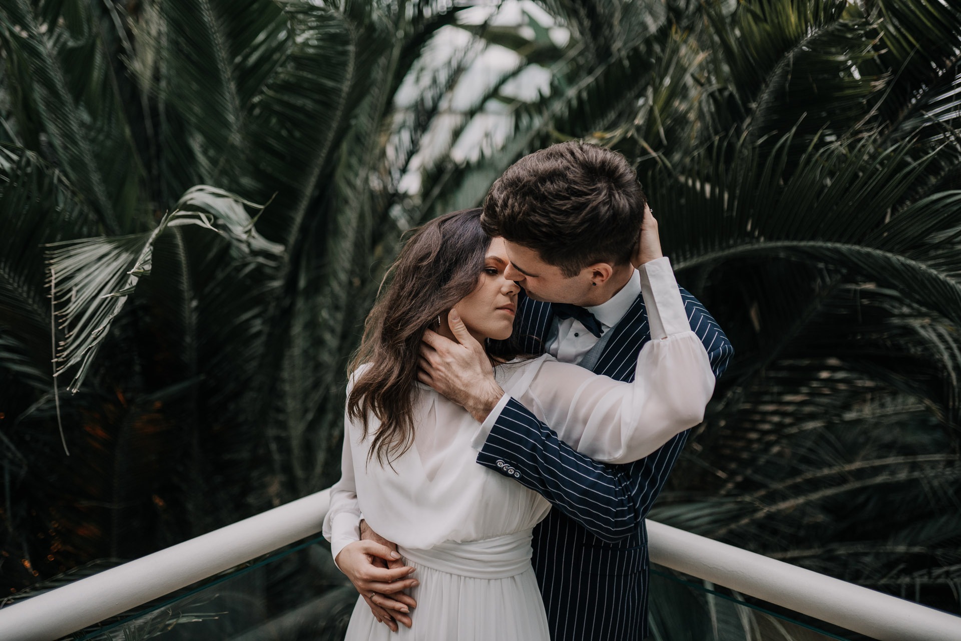 Para w eleganckich strojach, obejmująca się na tle egzotycznej roślinności w szklarni, mężczyzna w granatowym garniturze w prążki i muszce, kobieta w zwiewnej białej sukience.