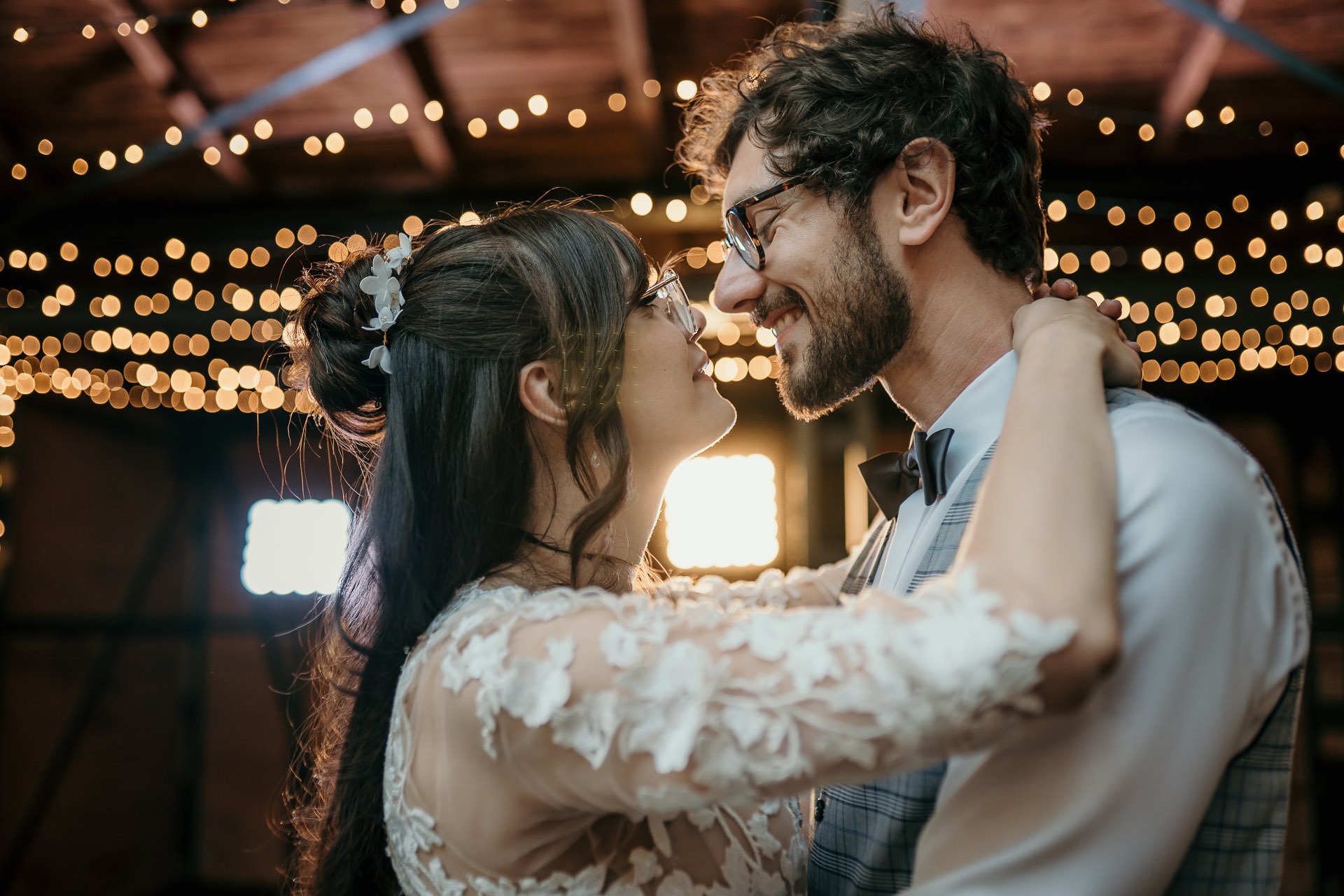 Uśmiechnięta para w okularach, obejmująca się twarzami w zbliżeniu, dekoracyjne lampki w tle.