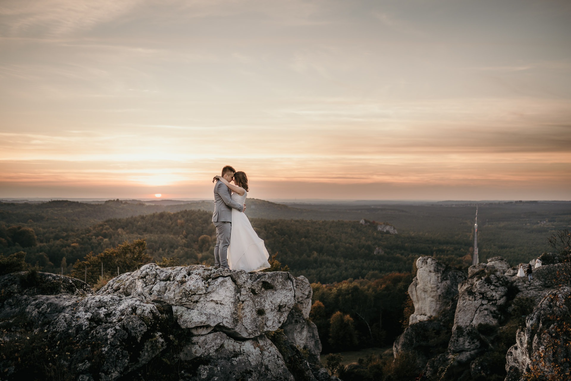 Para narzeczeńska w objęciach na skalistym wzgórzu o zachodzie słońca, malowniczy krajobraz leśny w tle.