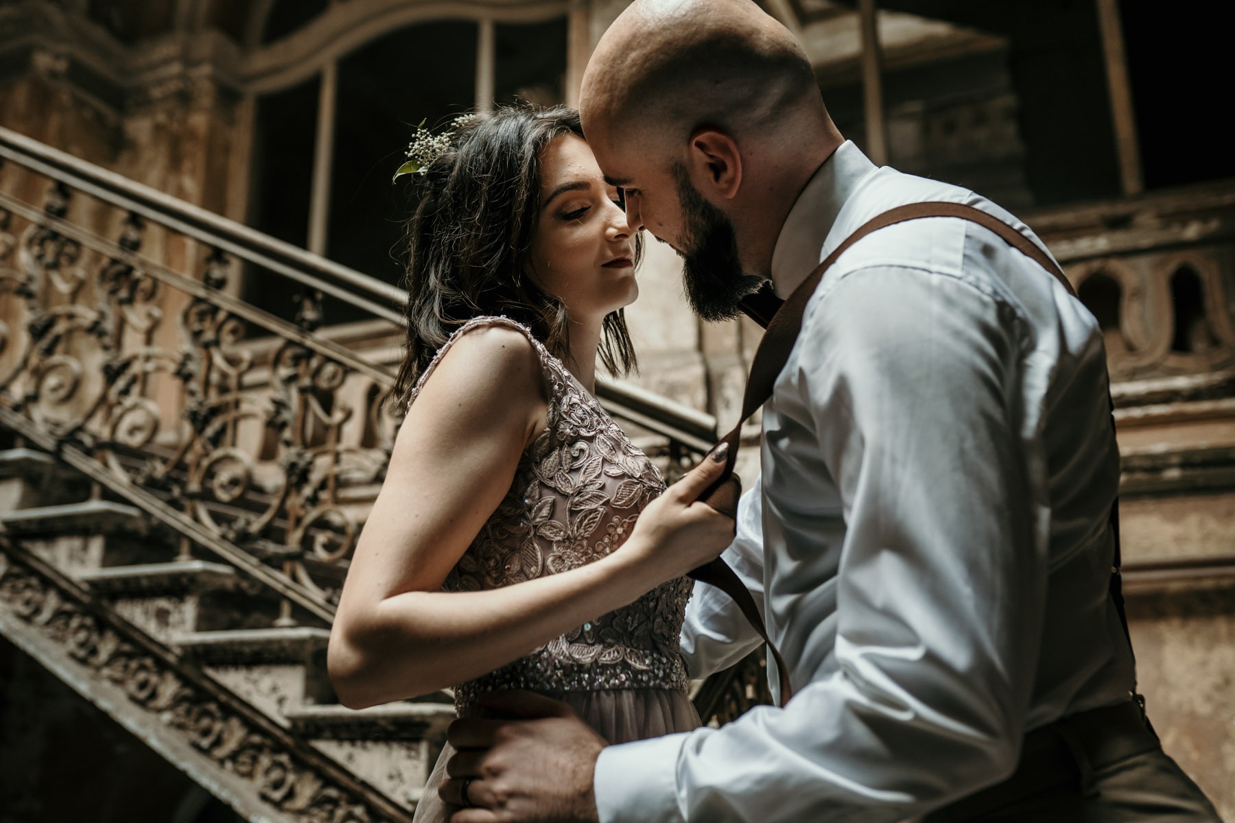Zakochana para w objęciach na tle zabytkowych schodów z kutą balustradą; ona w pudrowej sukience z koronką, on w białej koszuli z szelkami.