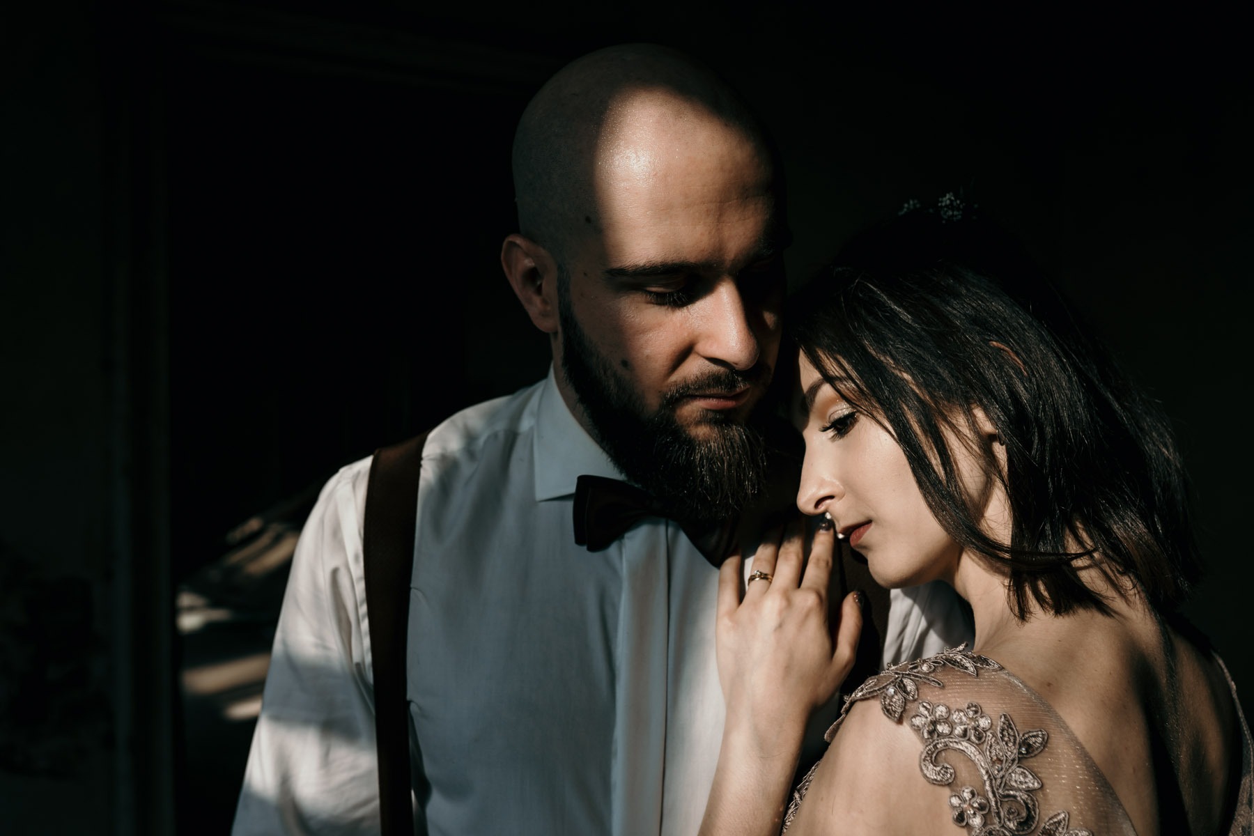 Para narzeczonych w zbliżeniu: mężczyzna z brodą w białej koszuli z muchą i szelkami, kobieta z ciemnymi włosami i delikatną sukienką z koronkowymi zdobieniami, uchwyceni w dramatycznym oświetleniu.