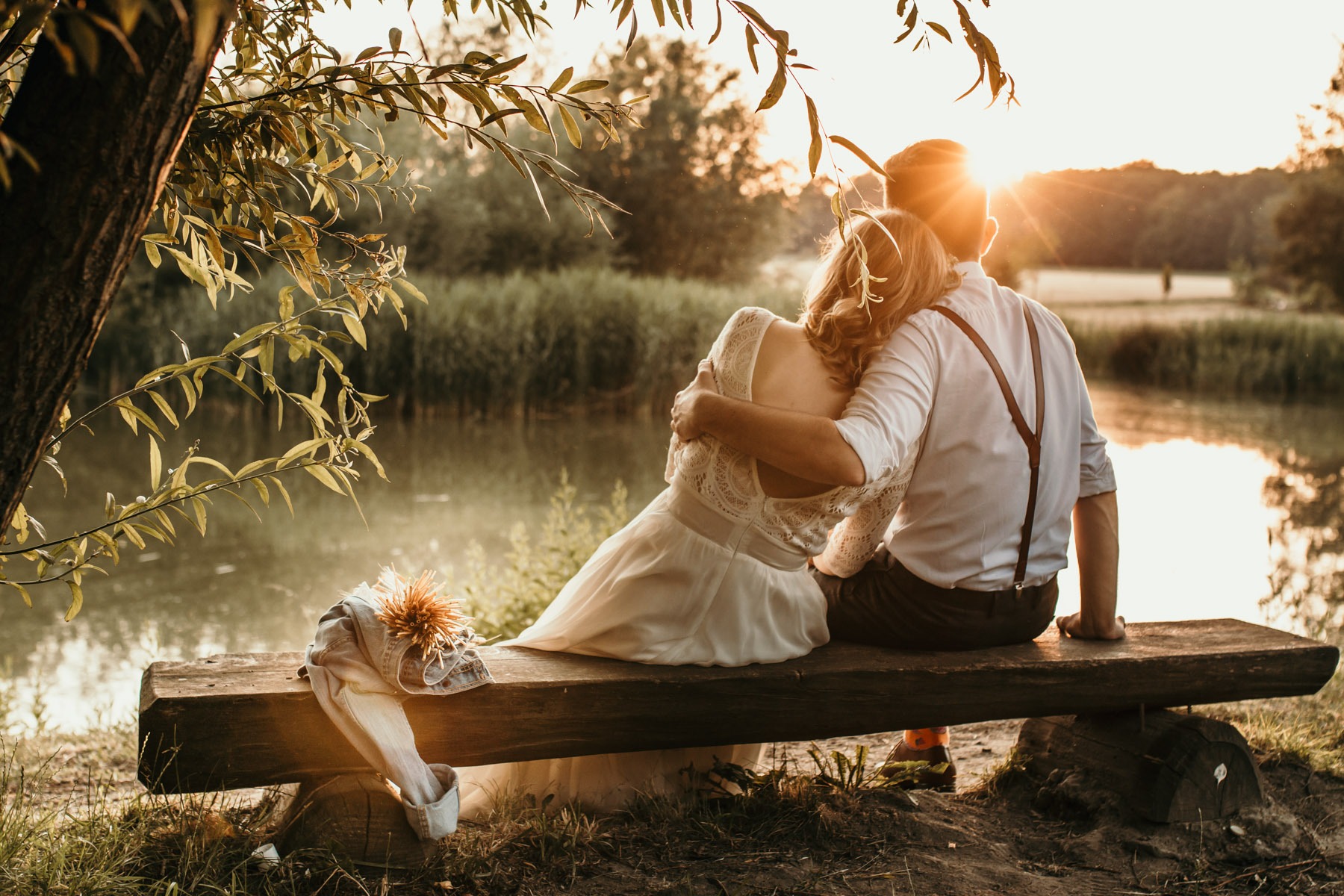 Para narzeczonych siedzi na drewnianej ławce nad jeziorem o zachodzie słońca, buty i bukiet leżą obok nich.