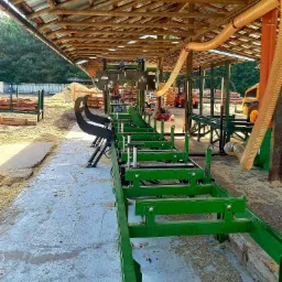 Widok na linię produkcyjną z zielonymi elementami konstrukcyjnymi w zadaszonym obiekcie, z widocznymi w tle stertami drewna i maszynami budowlanymi.