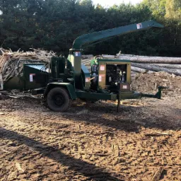 Zielona, mobilna rębarka do drewna na przyczepie, z stosem pociętych desek i pni drzew w tle, na błotnistym podłożu.