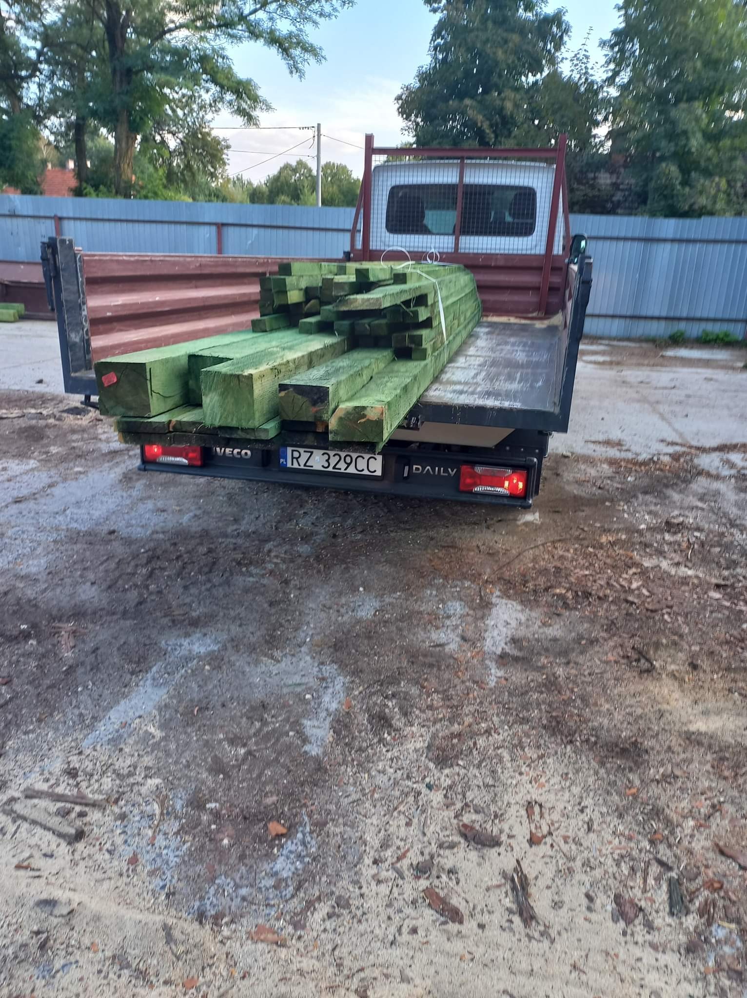 Ciężarówka Iveco Daily z zielonymi, impregnowanymi belkami drewnianymi na pace, widoczne tablice rejestracyjne RZ 329CC, teren placu budowy w tle.