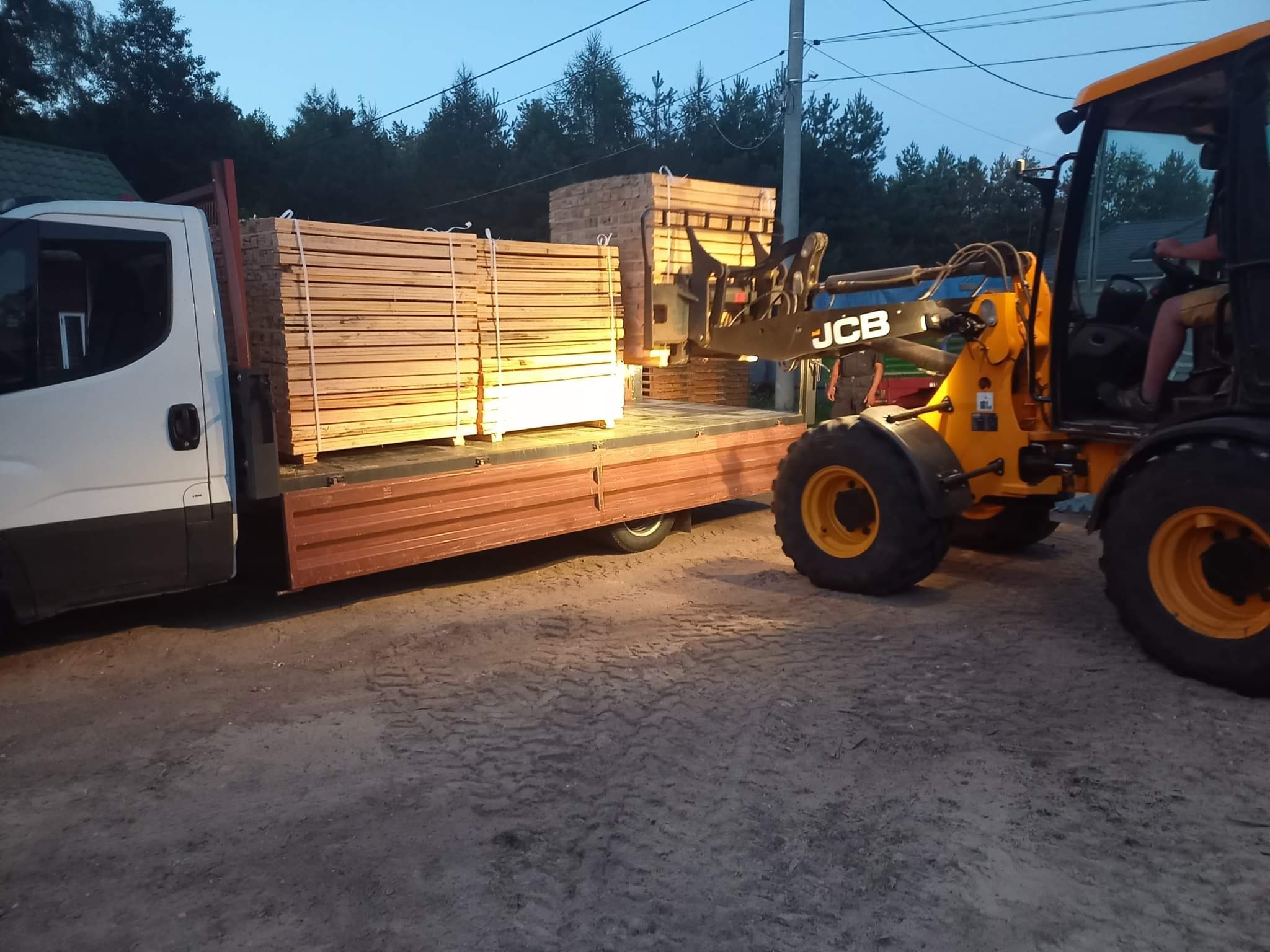 Żółta ładowarka JCB załadowuje palety z drewnem na samochód dostawczy w składzie drewna, widoczne ślady opon na piaszczystym podłożu, w tle drzewa.