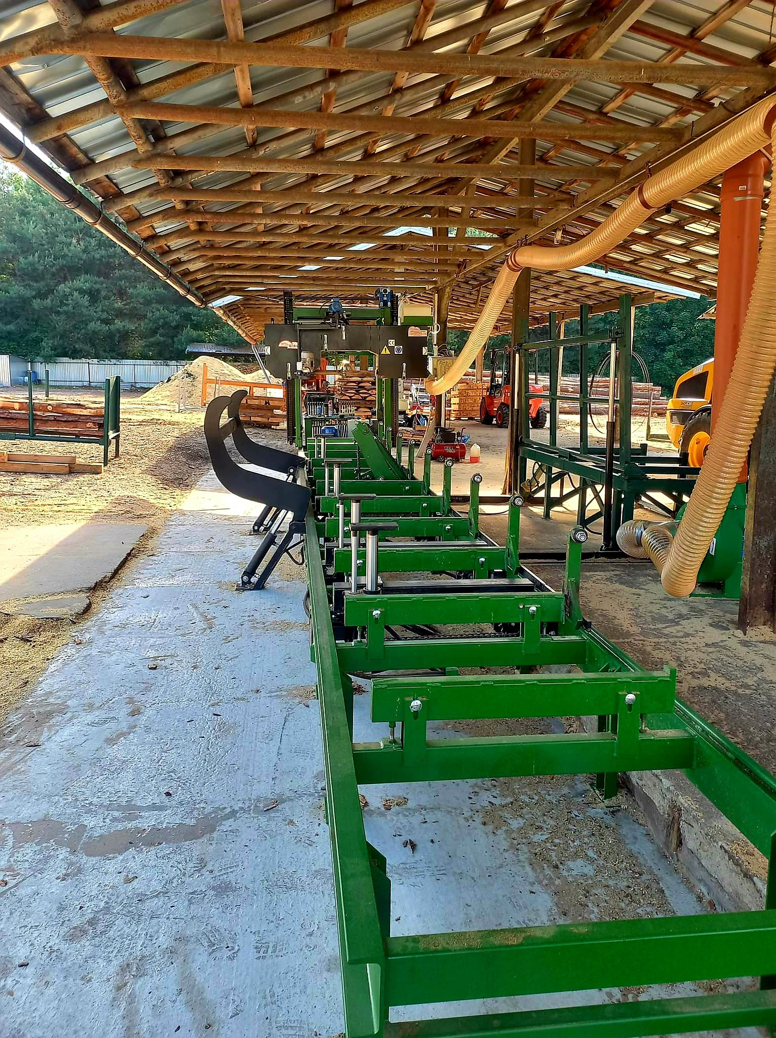 Widok na linię produkcyjną z zielonymi elementami konstrukcyjnymi w zadaszonym obiekcie, z widocznymi w tle stertami drewna i maszynami budowlanymi.