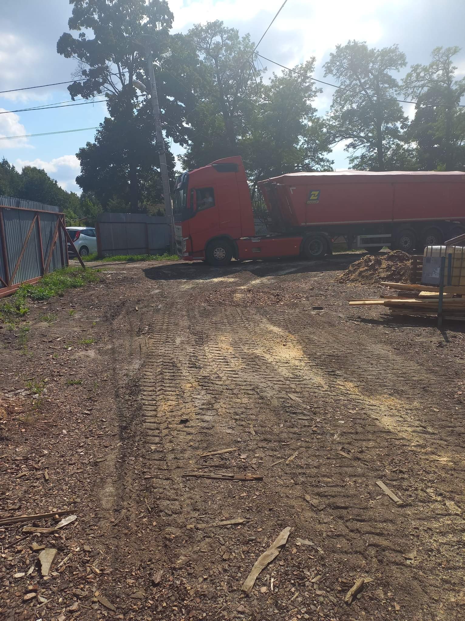 Czerwona ciężarówka z naczepą ZASŁAW na placu z widocznymi śladami opon i składowanym drewnem w tle, ogrodzenie po lewej.