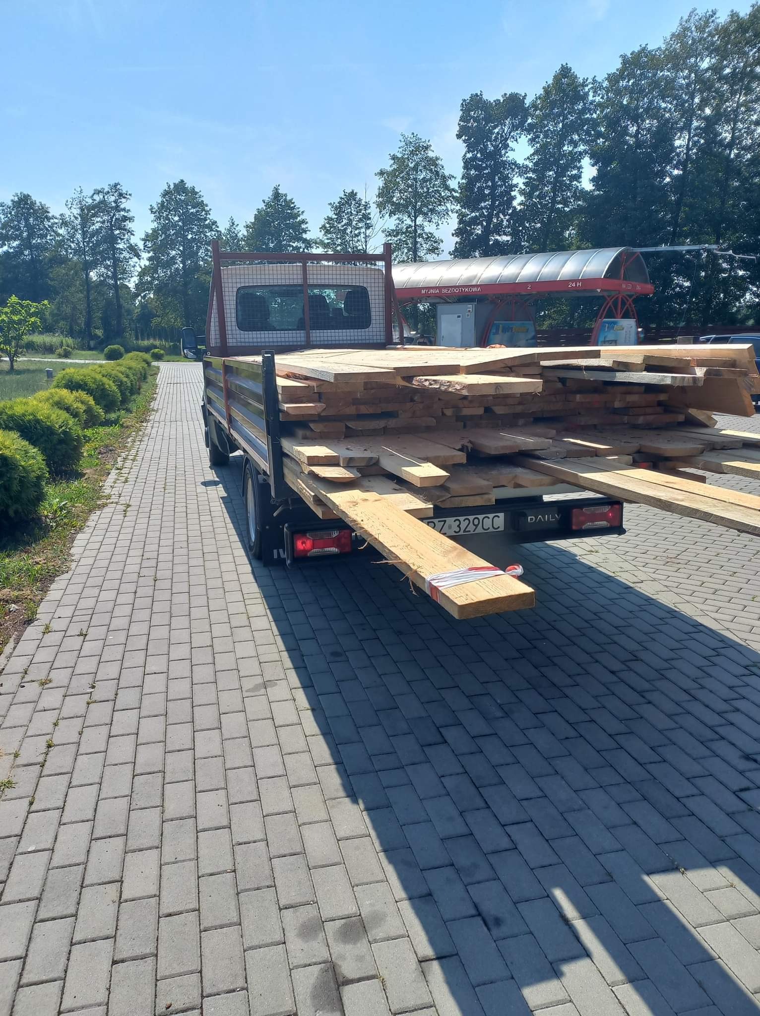 Ciężarówka z otwartą skrzynią ładunkową wypełnioną drewnem, zaparkowana na brukowanym placu w słoneczny dzień, w tle myjnia bezdotykowa.