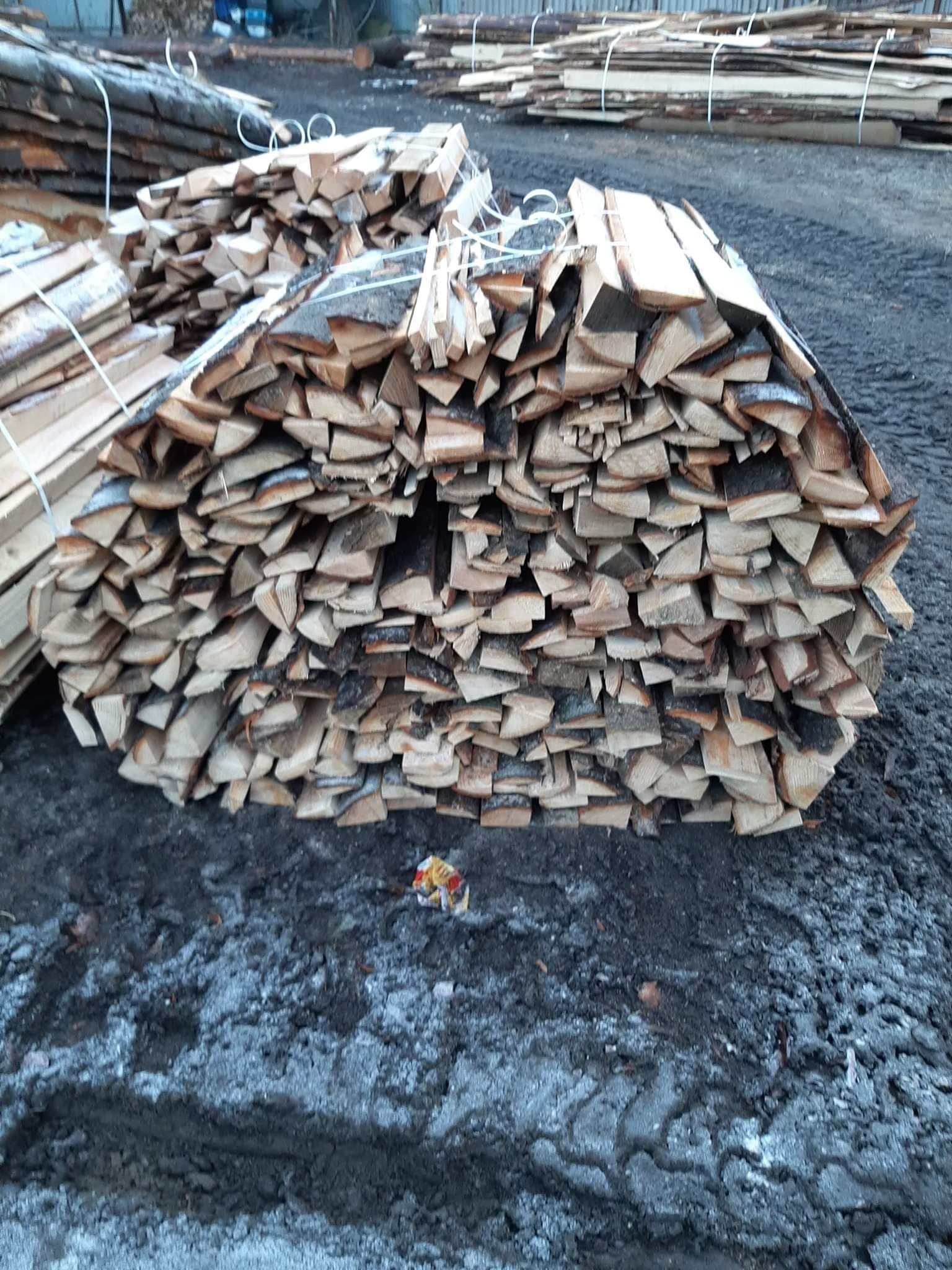 Stos pociętego drewna opałowego, zabezpieczonego sznurkiem, ułożony na ziemi z resztkami śniegu, w tle widoczne kolejne stosy drewna.