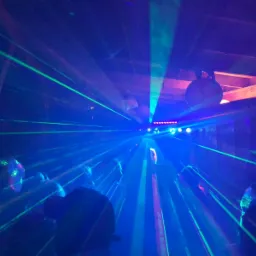 Intensywne niebieskie i zielone lasery przecinają ciemne pomieszczenie z widocznymi elementami konstrukcyjnymi sufitu i dyskotekową kulą. Reflektory na suficie tworzą punkt centralny.