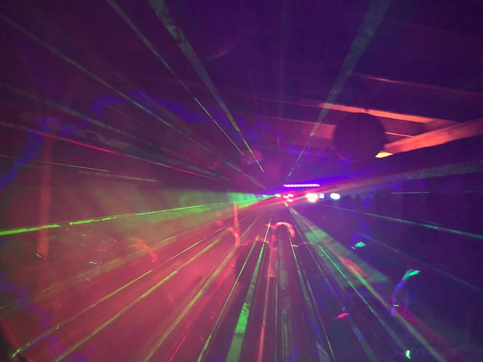 Wiązki zielonych, czerwonych i niebieskich świateł laserowych przecinają ciemne pomieszczenie podczas imprezy. Widoczni ludzie w tle.