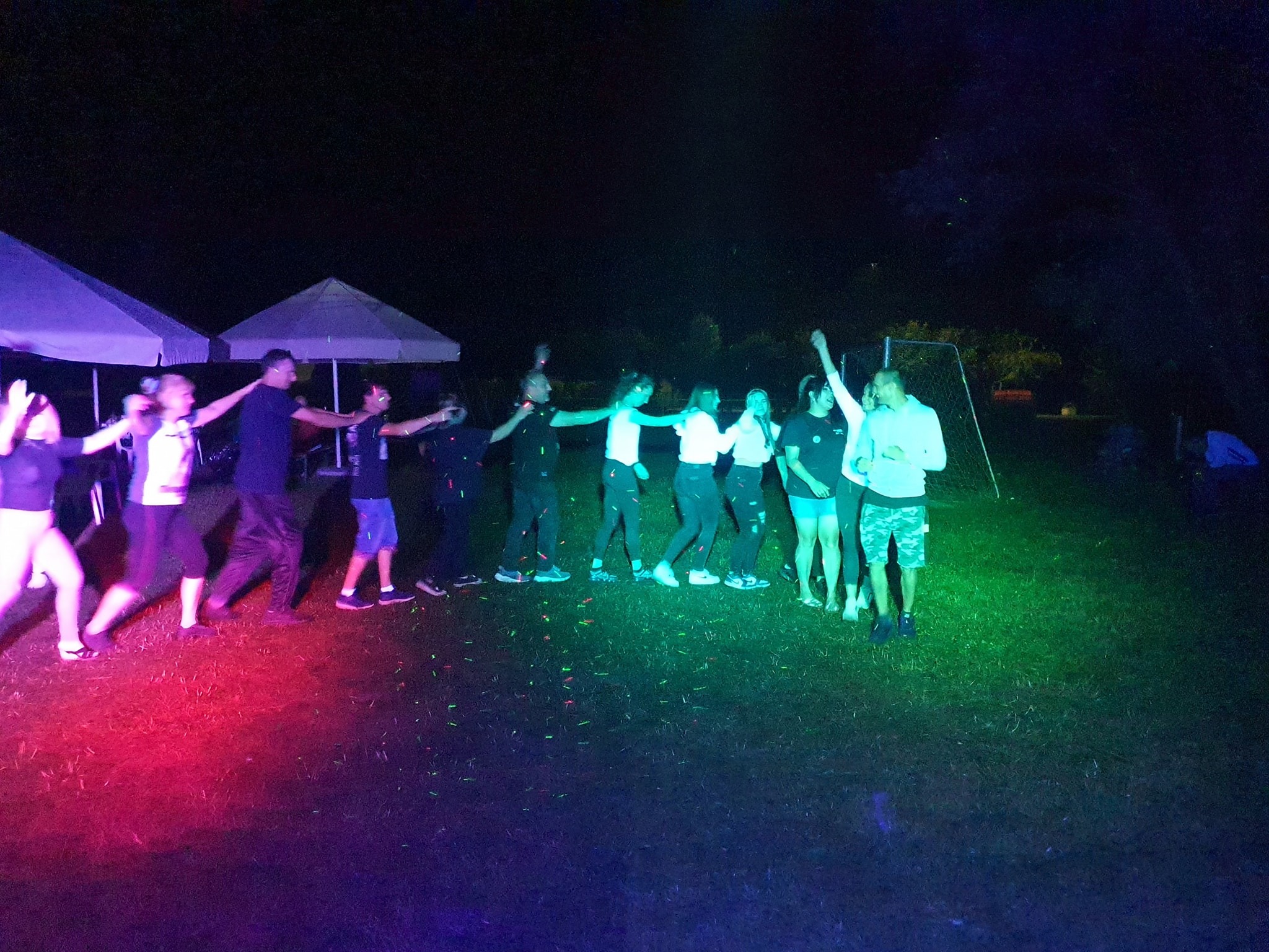 Grupa osób trzymających się za ręce, tańczących na trawie w nocy, oświetlonych czerwonym i zielonym światłem, widoczne namioty w tle.