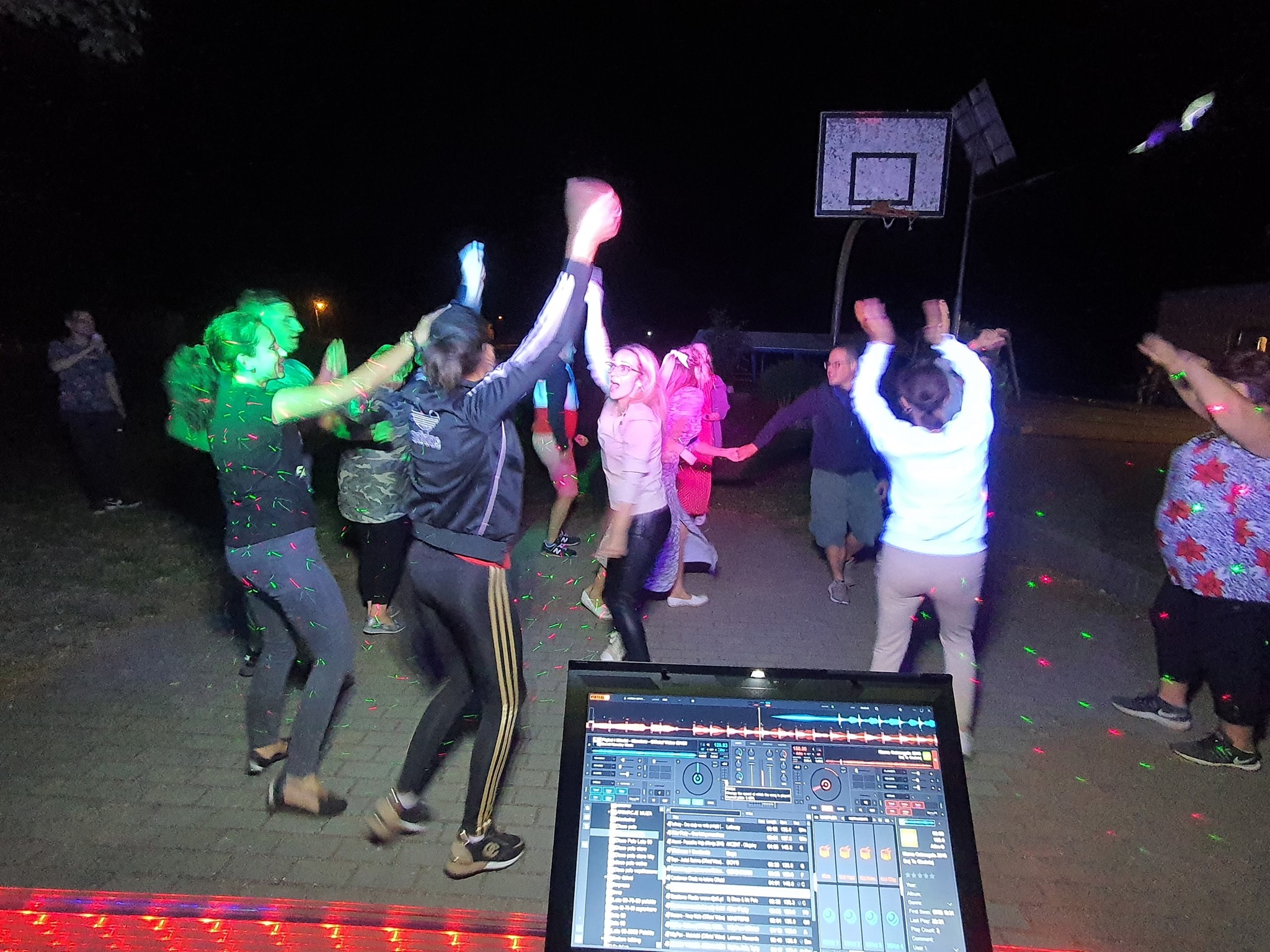 DJ puszcza muzykę na imprezie plenerowej podświetlonej kolorowymi światłami, w tle tańczący ludzie i kosz do koszykówki.
