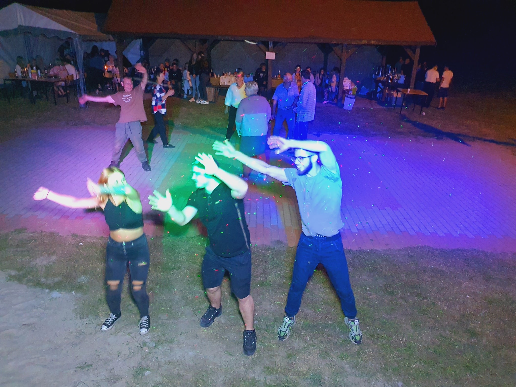 Grupa osób tańczy na zewnątrz w wieczornym oświetleniu dyskotekowym, z widoczną sceną w tle i publicznością.