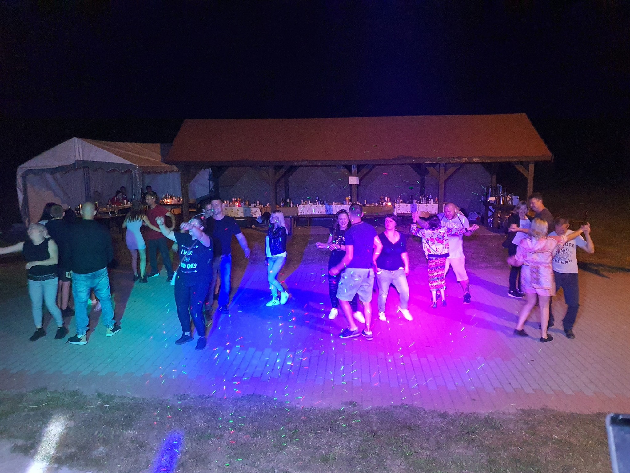 Grupa osób tańczących na zewnątrz pod zadaszeniem, oświetlonych kolorowymi światłami, z widocznym cateringiem na stołach w tle.