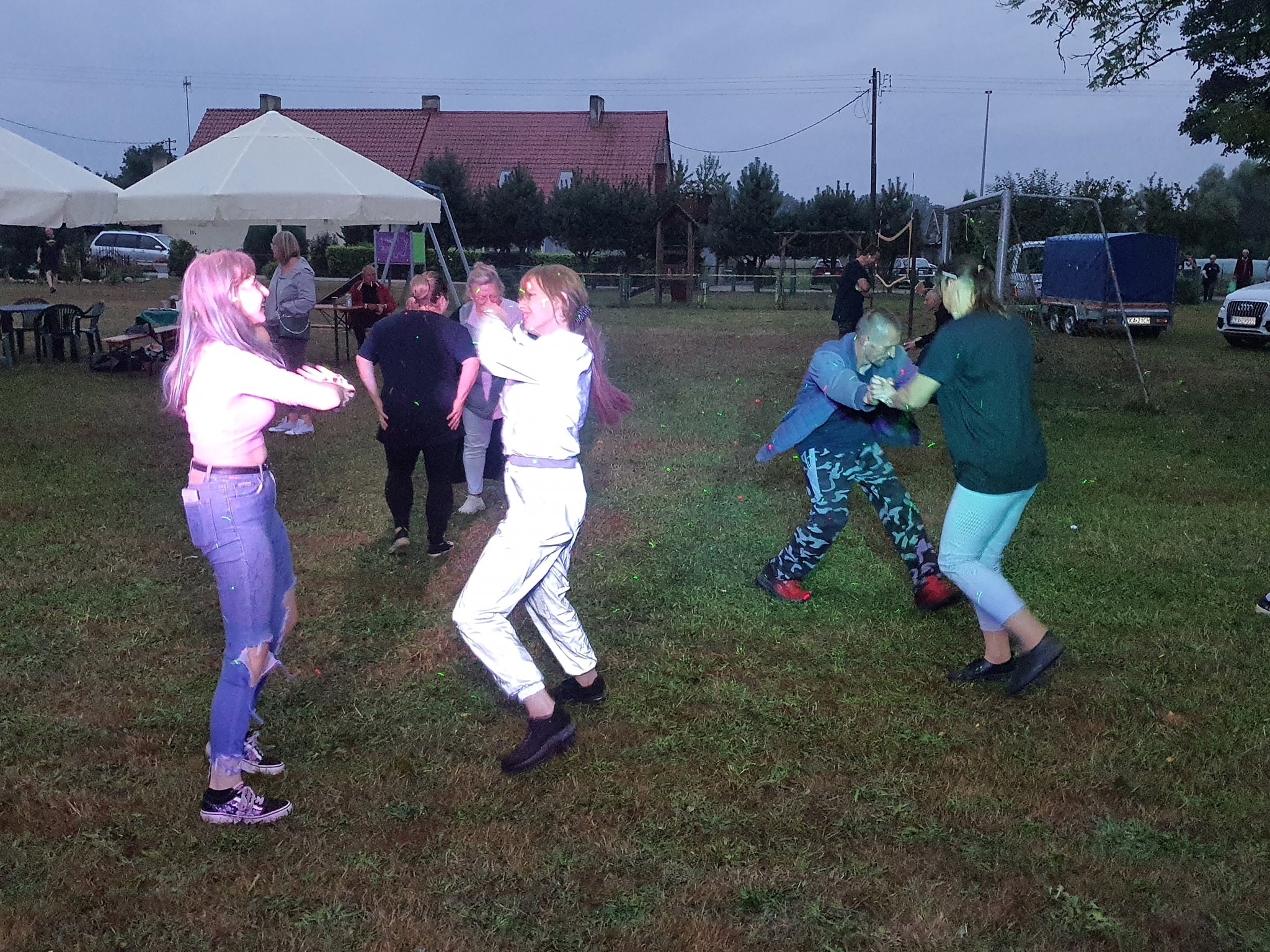 Uczestnicy imprezy plenerowej tańczą na trawie, widoczne namioty i budynek w tle, oświetlenie sceniczne z efektem zielonych kropek.