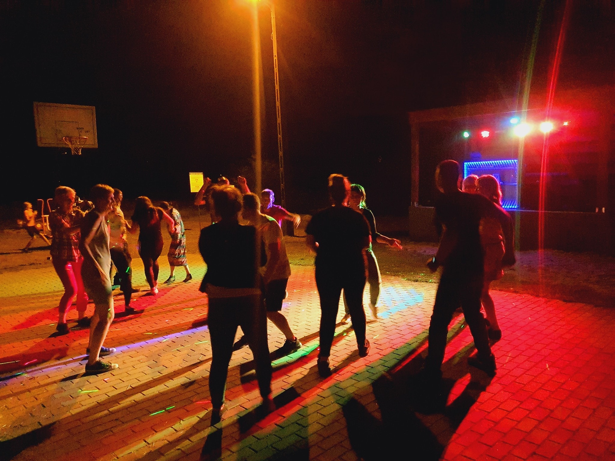 Nocna impreza plenerowa z tańczącymi osobami oświetlonymi kolorowymi światłami, w tle scena z podświetleniem LED i kosz do koszykówki